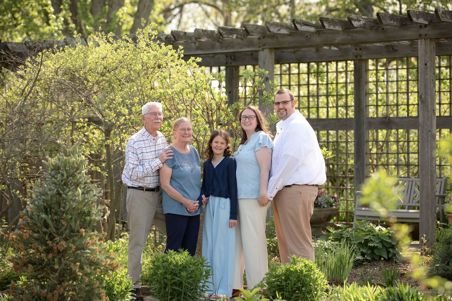 Family of five smiling together in a lush garden setting beneath a wooden pergola on a sunny spring day.