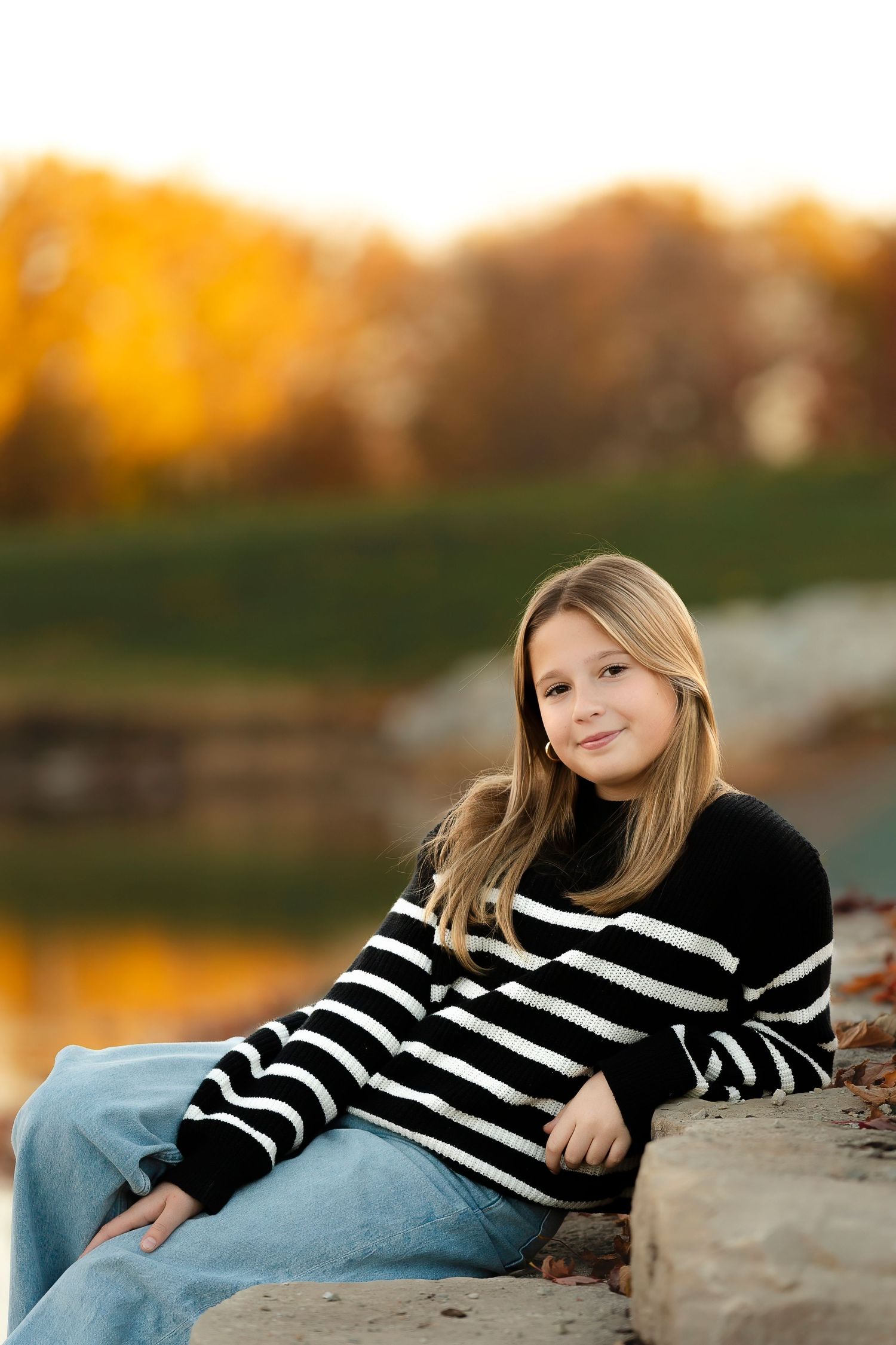 Person in black and white striped sweater sits on stone wall by lake during autumn sunset.