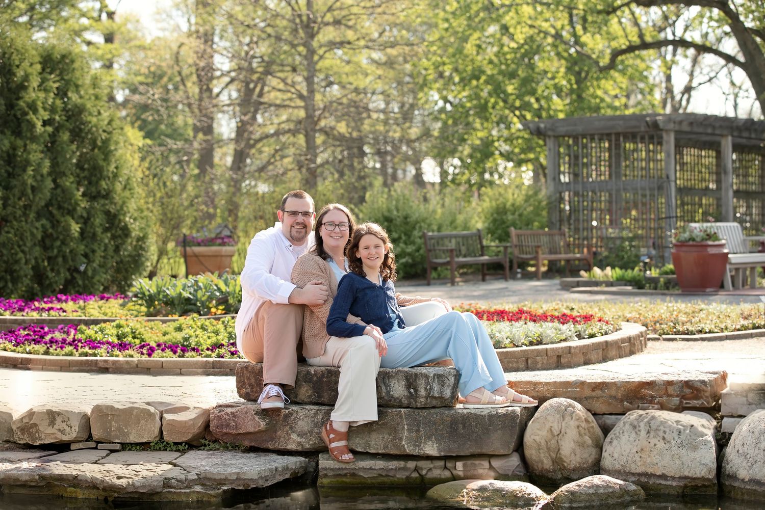 Family of three smiling together on stone steps near a garden pond surrounded by colorful flowers.