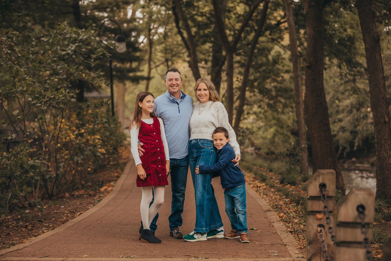 Family of four smiling together on a tree-lined path in autumn, parents with young daughter in red dress and son.