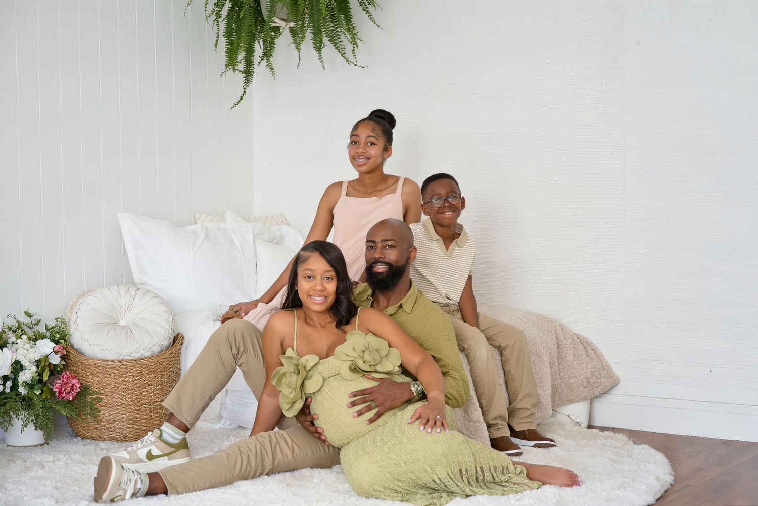 A joyful Black family of four poses together in a bright white room, wearing coordinated green and beige outfits.