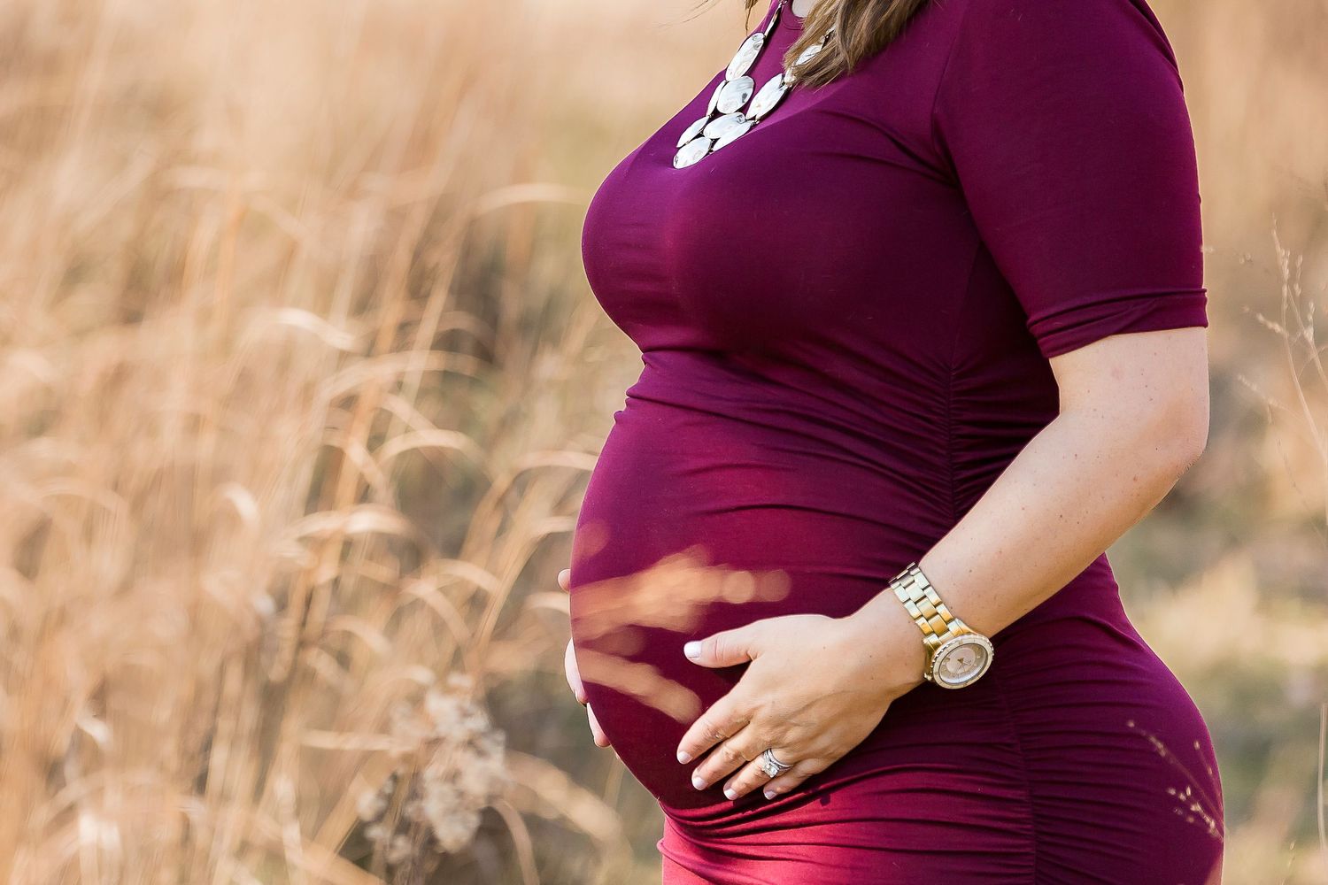 A pregnant woman in a burgundy dress cradles her belly while standing in a golden wheat field.
