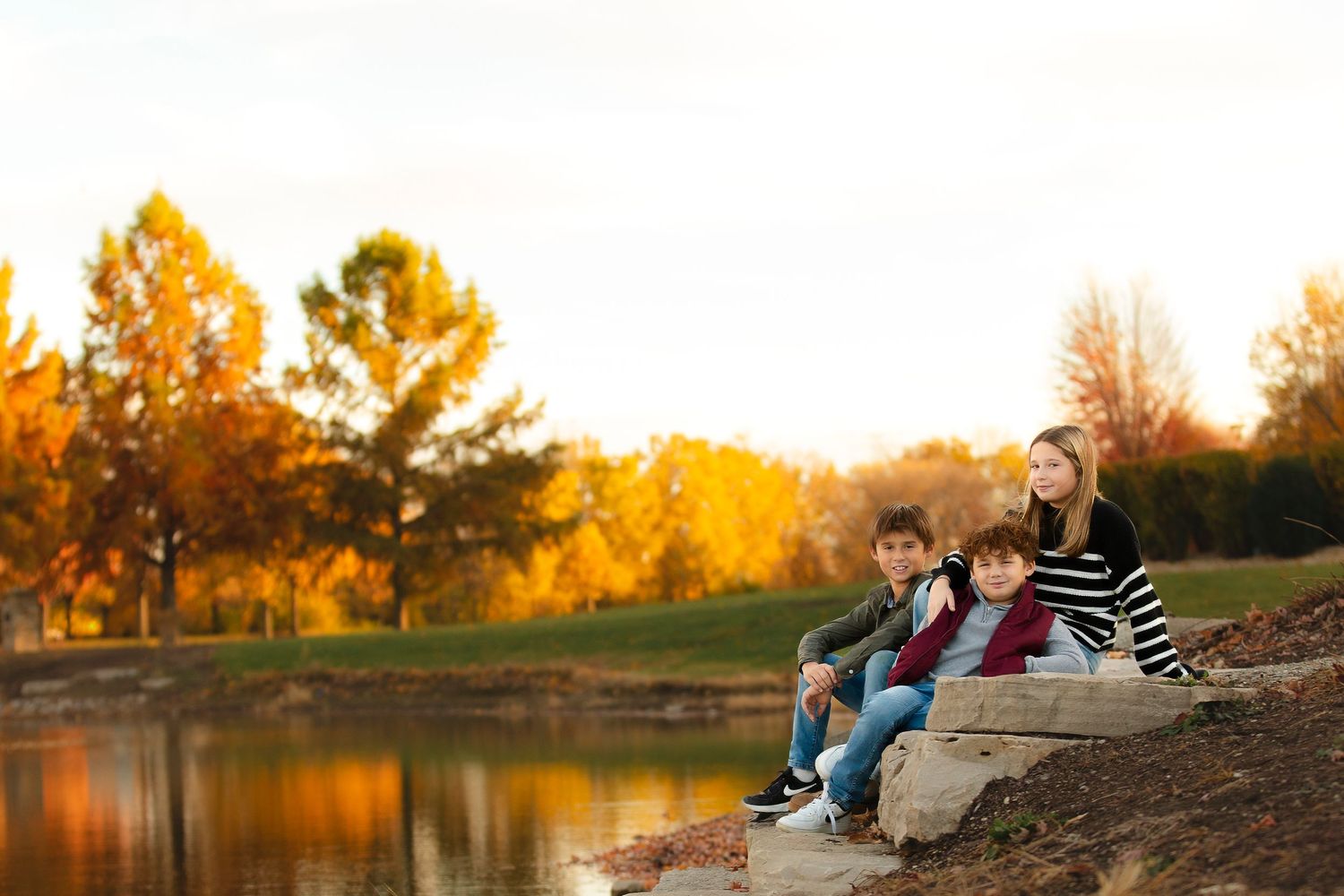 People sit together on a riverbank at sunset while golden autumn trees reflect in the calm water.