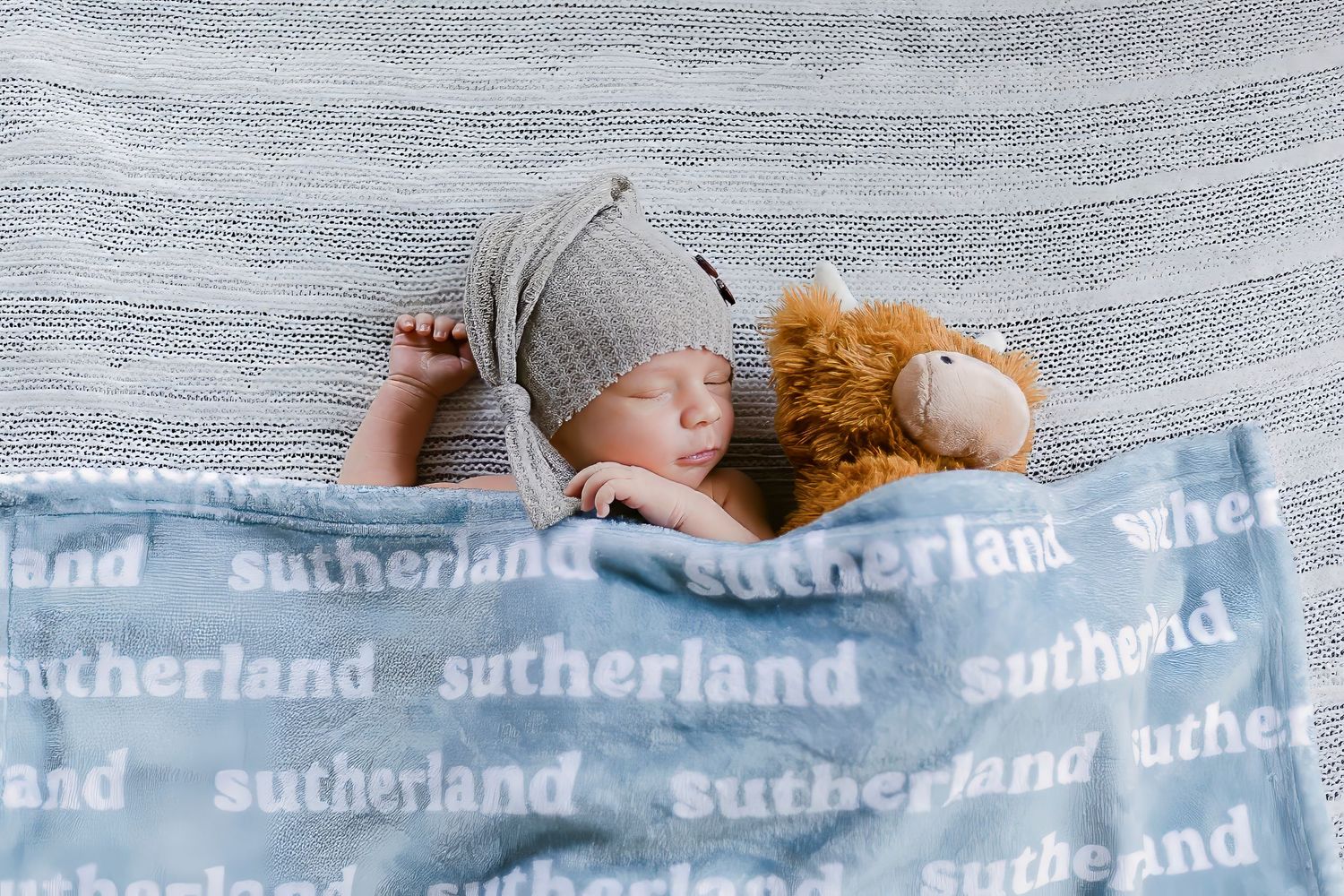 A sleeping baby in a gray knit hat cuddles with a brown teddy bear on a blue blanket with text pattern.