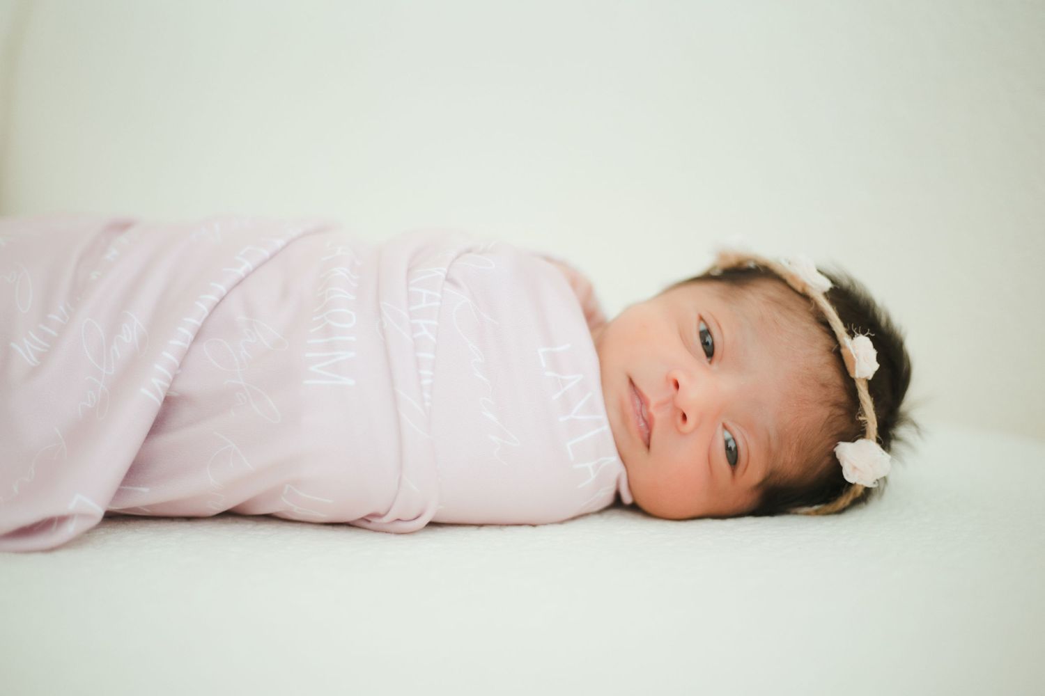 Newborn baby girl swaddled in a soft pink blanket wearing a white floral headband, lying on a white background.