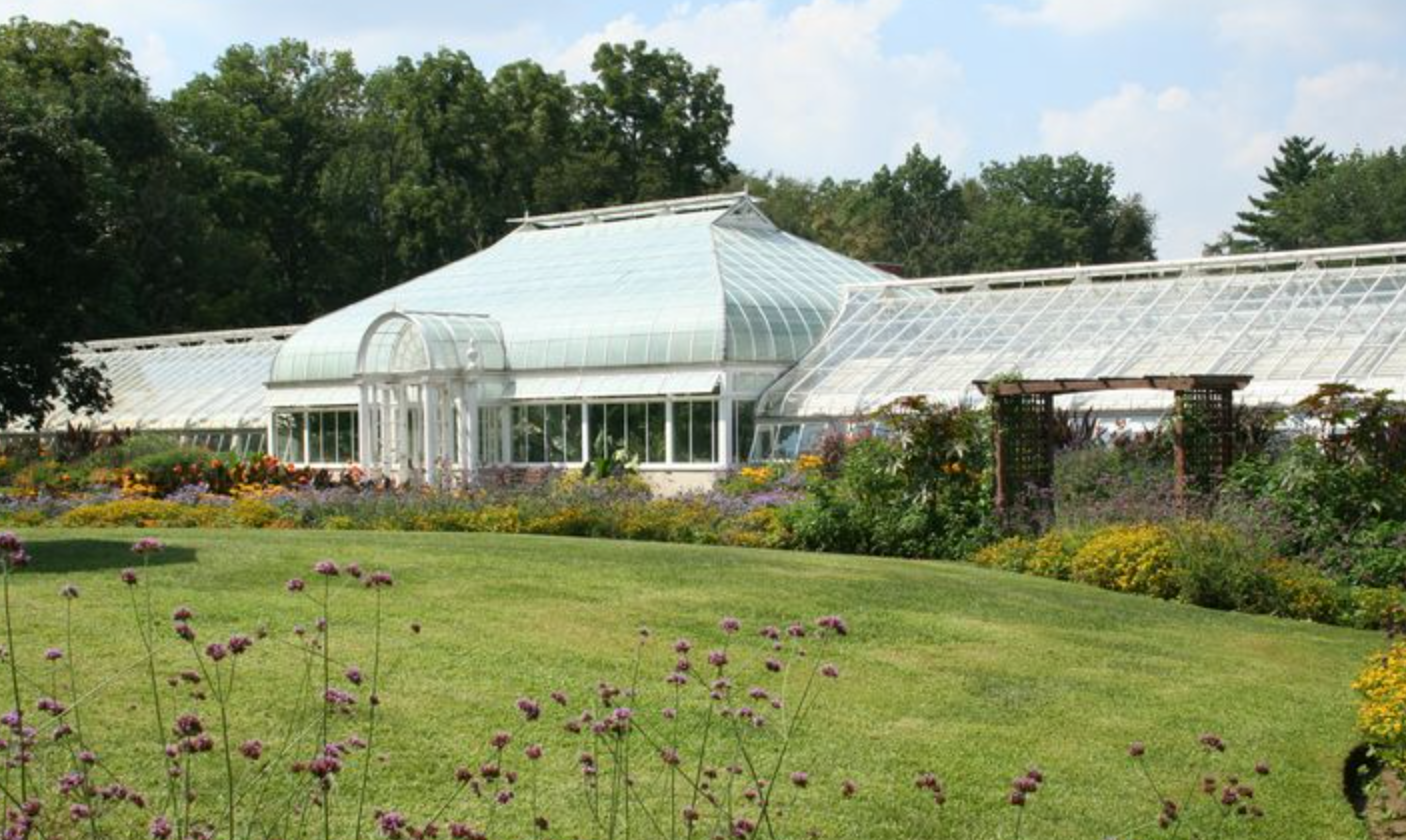 Lush botanical garden with large glass greenhouse, colorful flower borders, and manicured lawn on a sunny day.