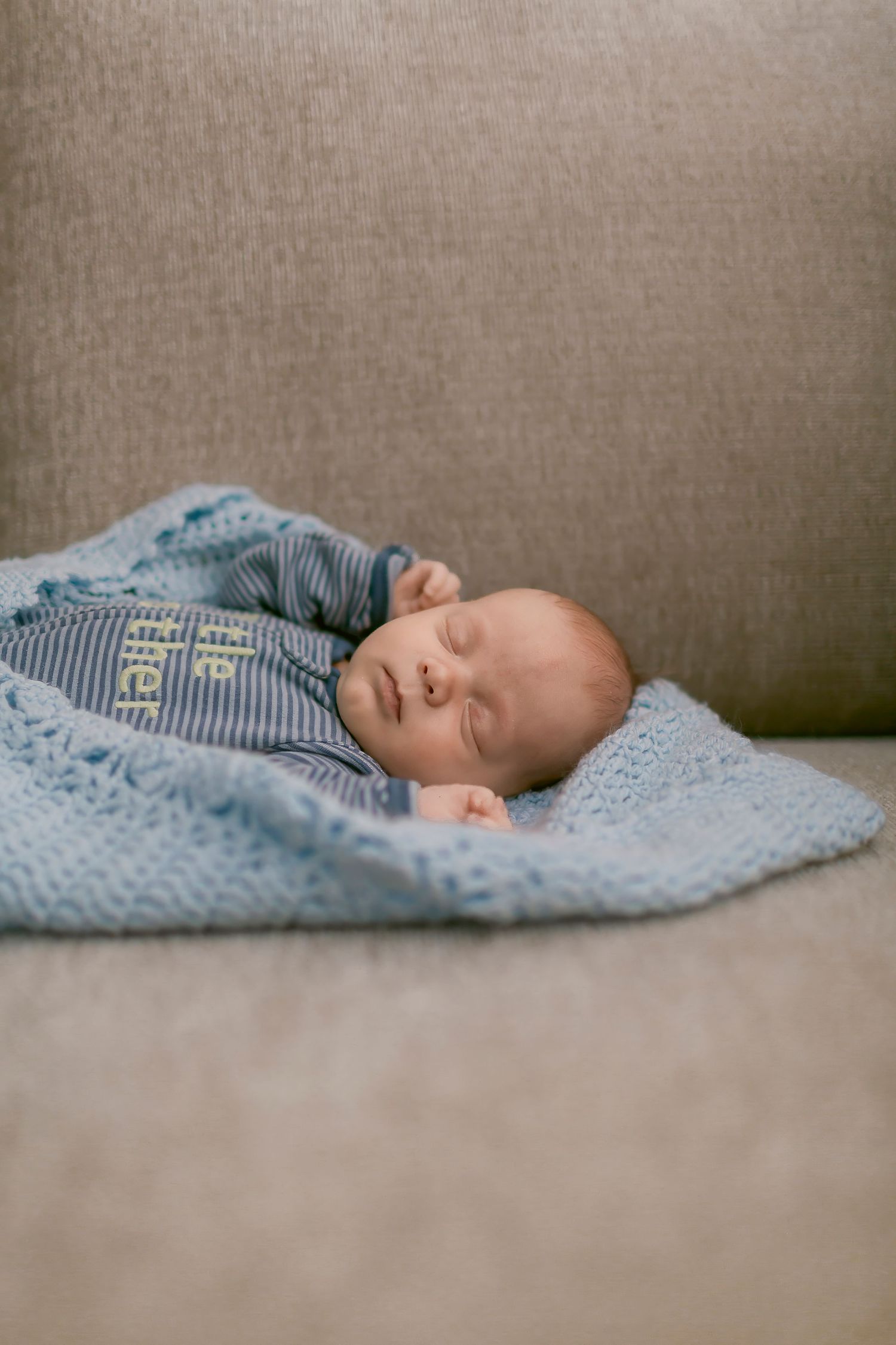 Newborn baby sleeping peacefully on a gray sofa, wrapped in a soft blue knit blanket wearing a striped blue outfit.