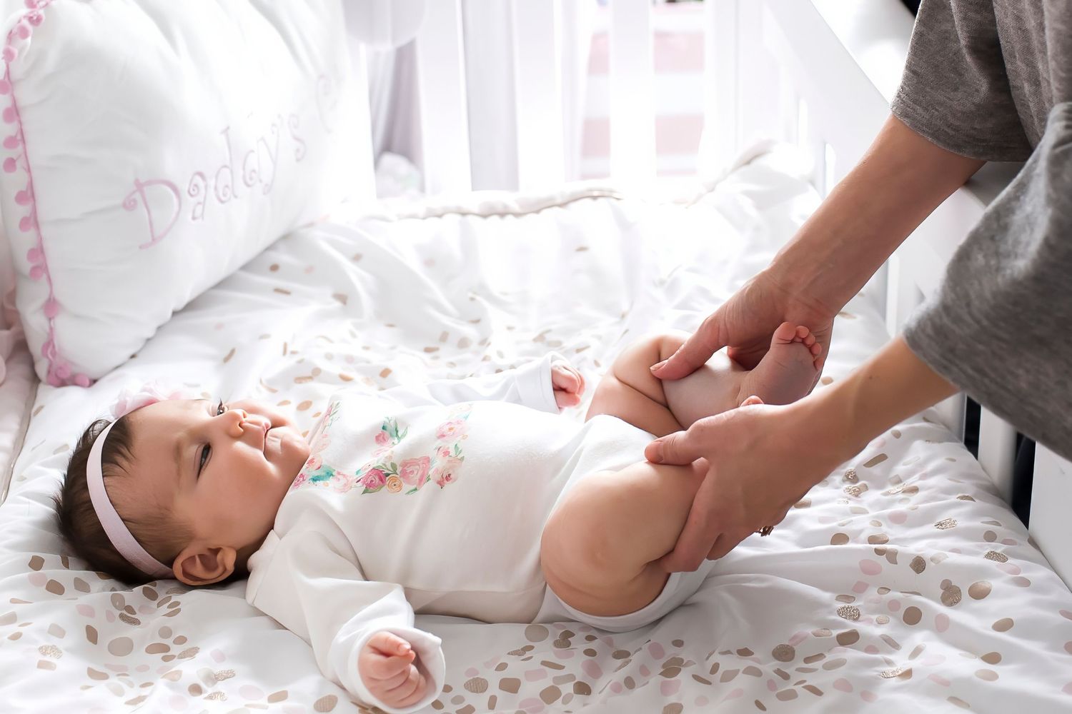 A newborn baby lies on white bedding while being gently touched by caring hands during a photography session.