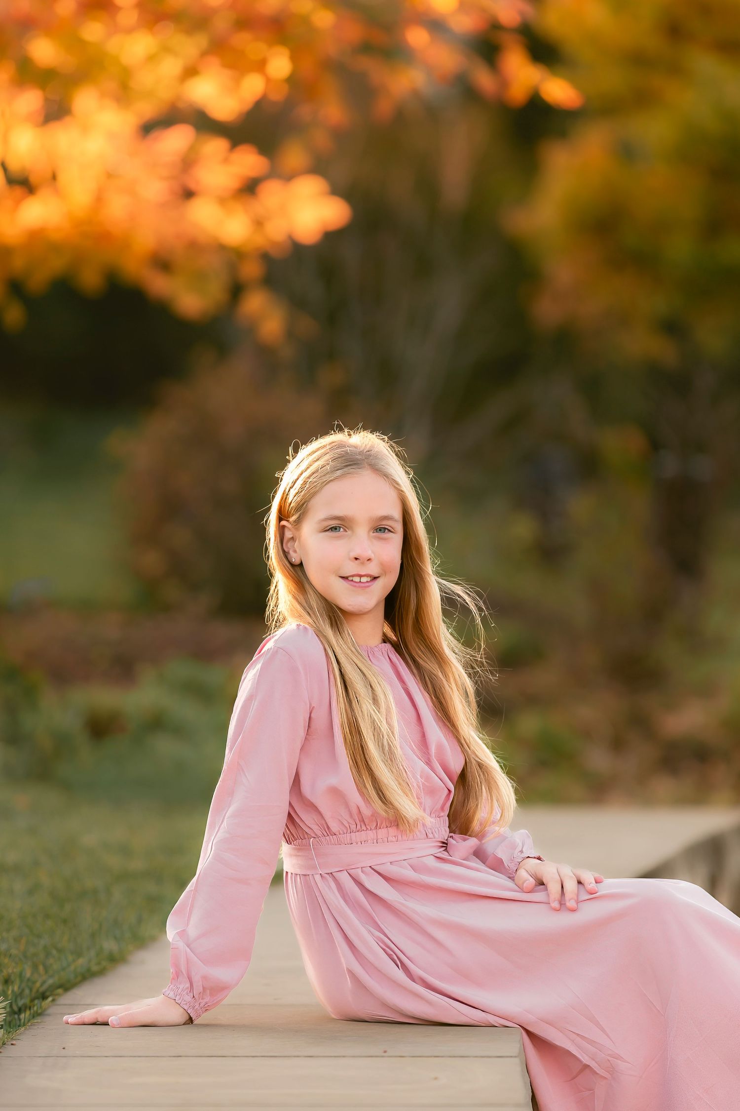 A child in a pink dress lies on a wooden surface during autumn with orange foliage in the background.