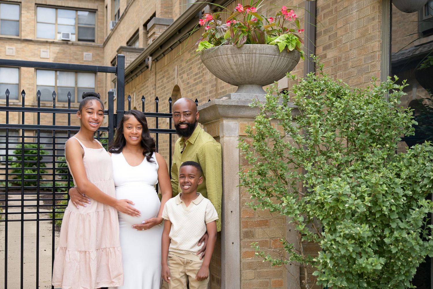 A happy family of four poses outdoors, with a pregnant woman in white, beside brick building with lush greenery.