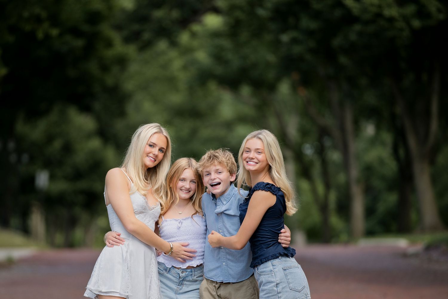 Four blonde siblings smile and hug outdoors on a tree-lined path, including two teenage girls, a young girl, and a boy.