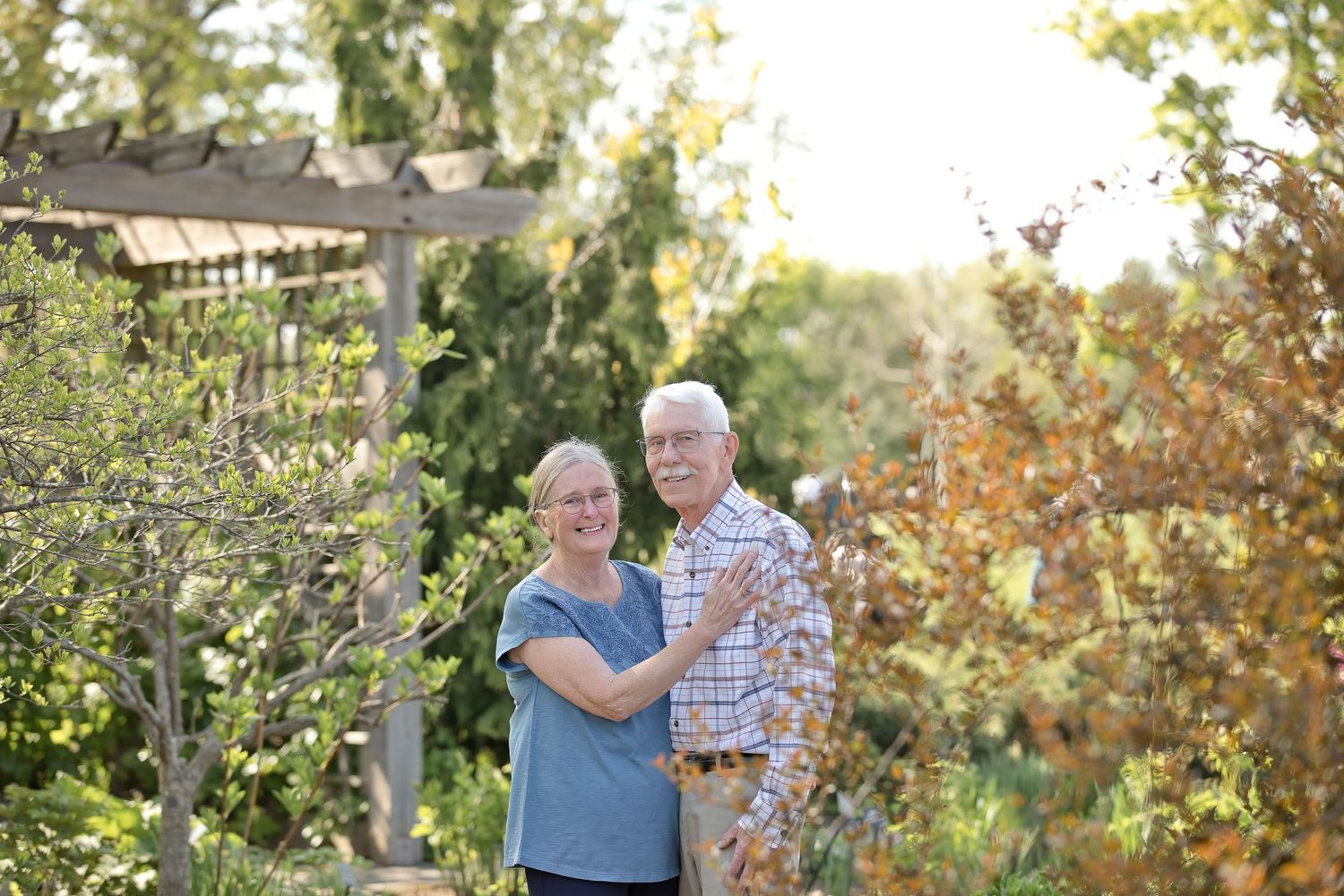 Elderly couple embracing and smiling in a lush garden with pergola and colorful autumn foliage on a sunny day.