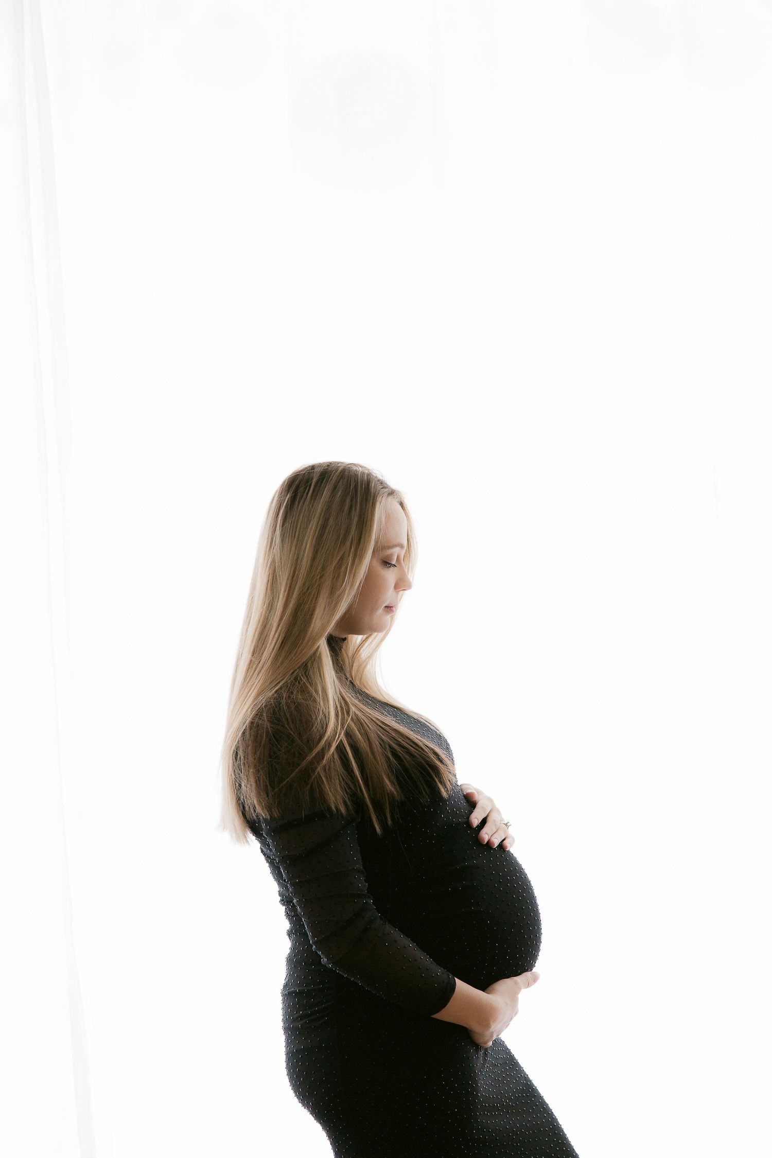 Pregnant woman in black dress cradling her baby bump, standing in profile against a bright white background.