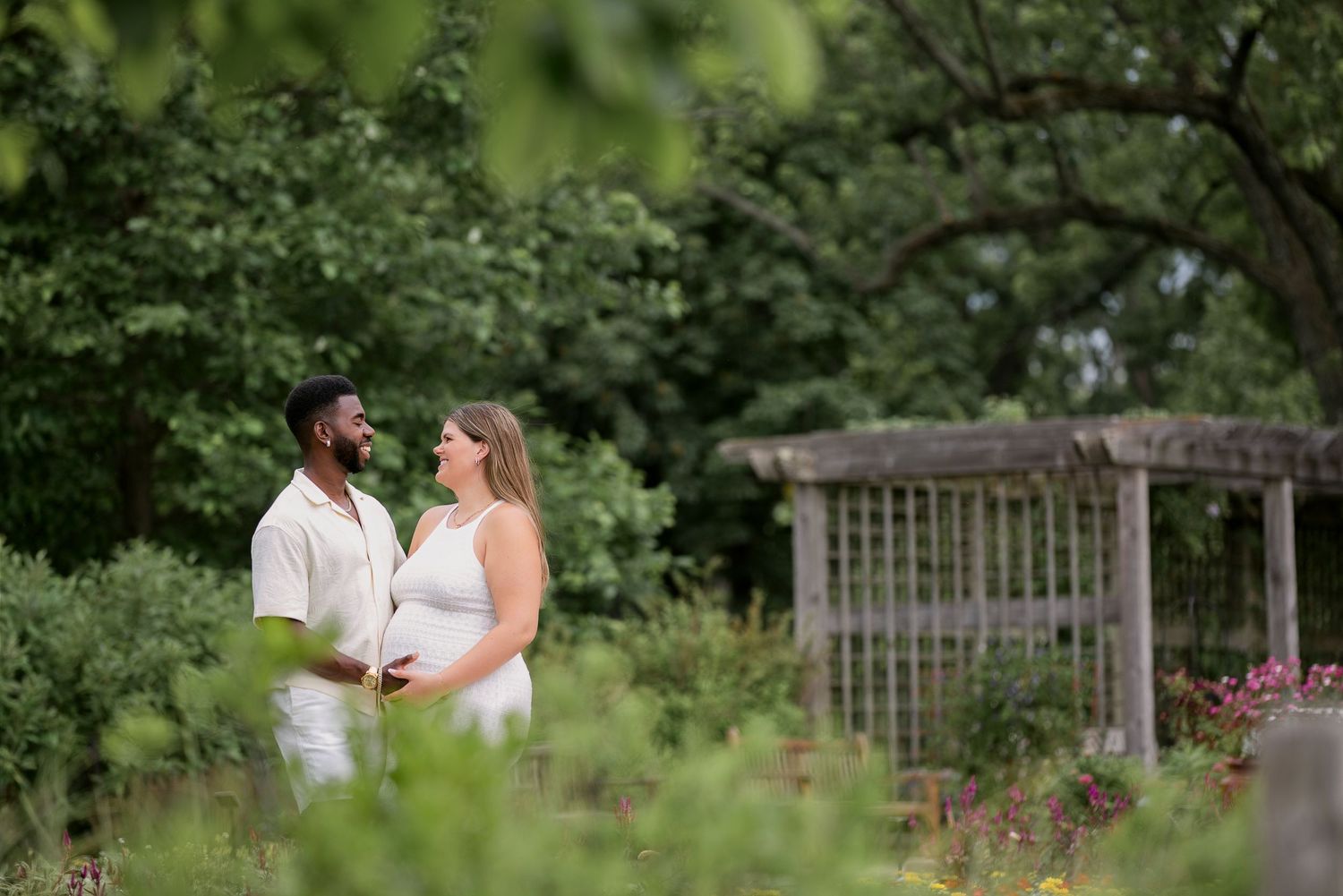 Expecting couple in white outfits sharing a tender moment in a lush green garden setting.
