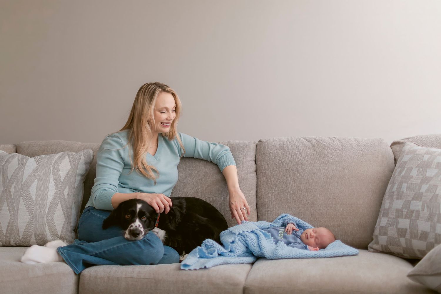 Woman smiling on gray sofa with black and white dog and sleeping newborn baby wrapped in blue blanket.