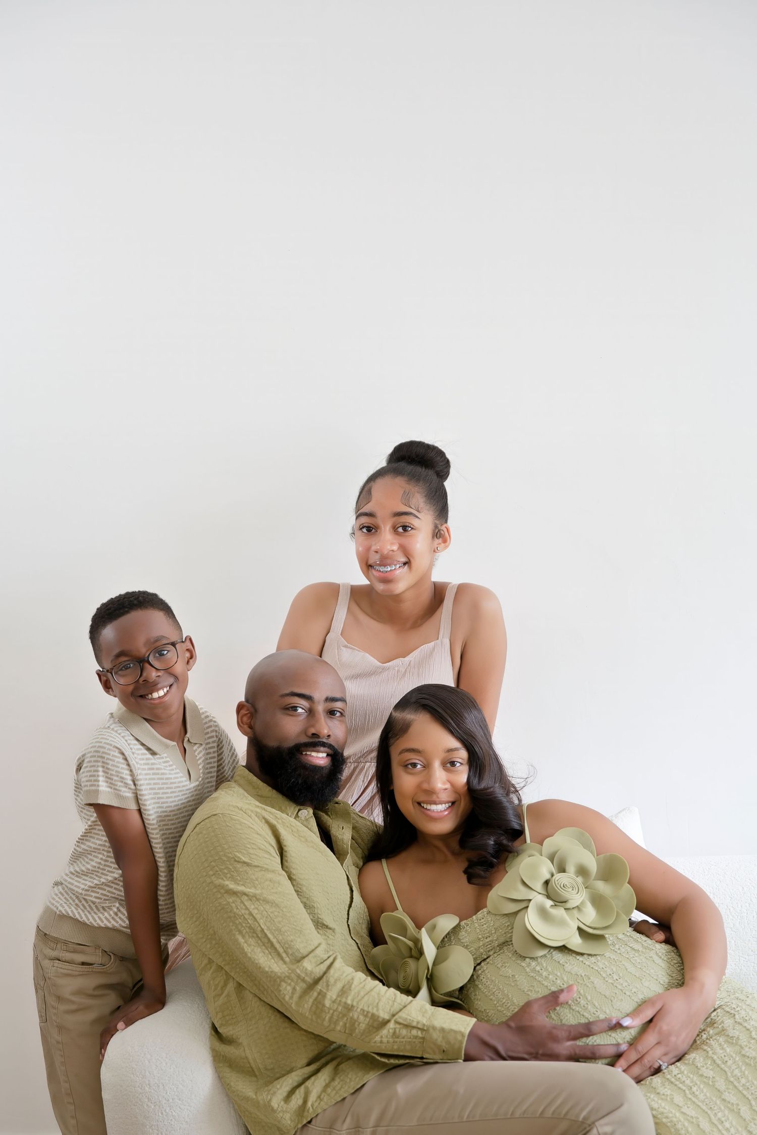 A joyful family of four poses together, with a pregnant woman in a green floral dress, a bearded man, and two children.