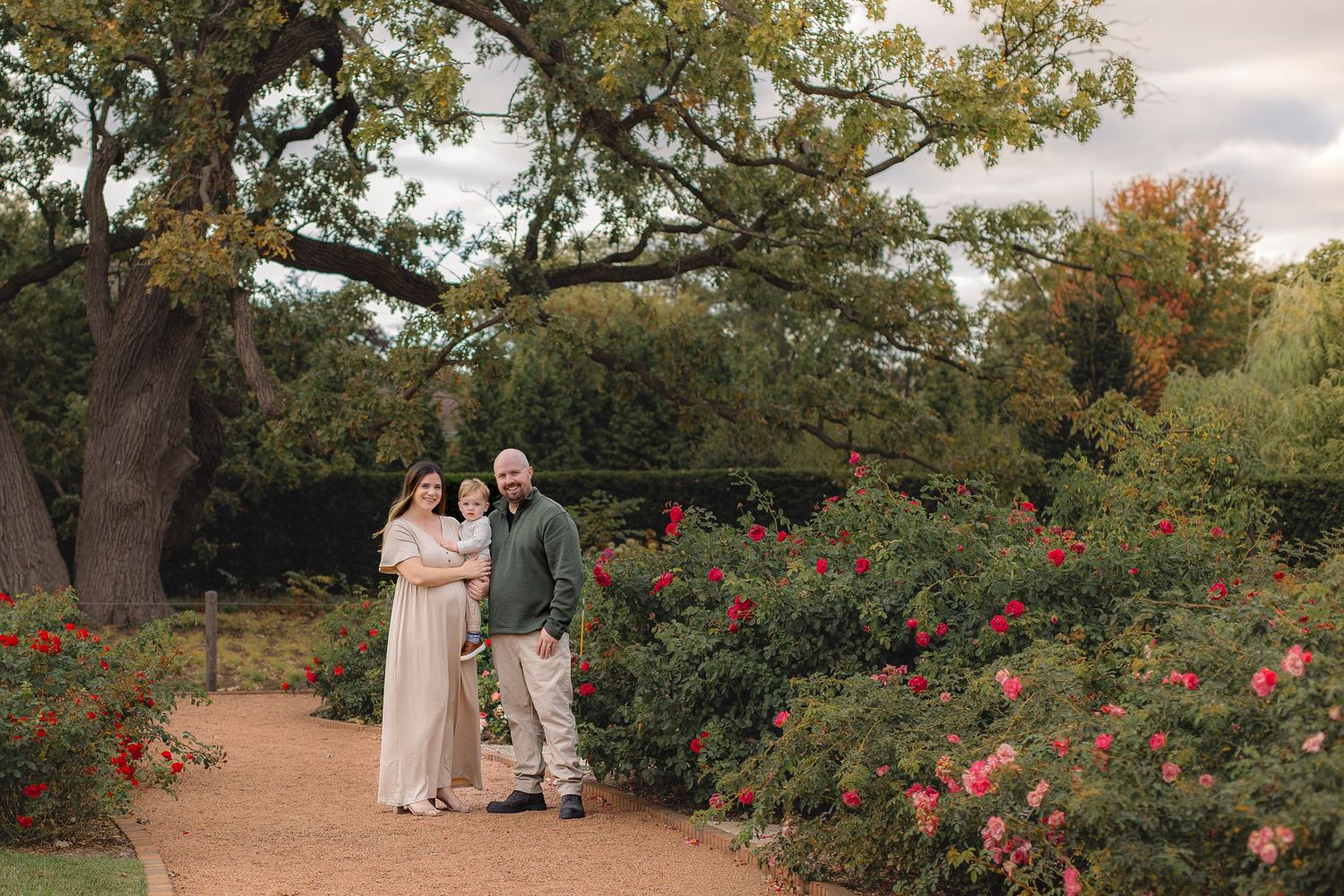 Two people share a romantic moment in a garden path surrounded by blooming rose bushes and trees on an autumn day.