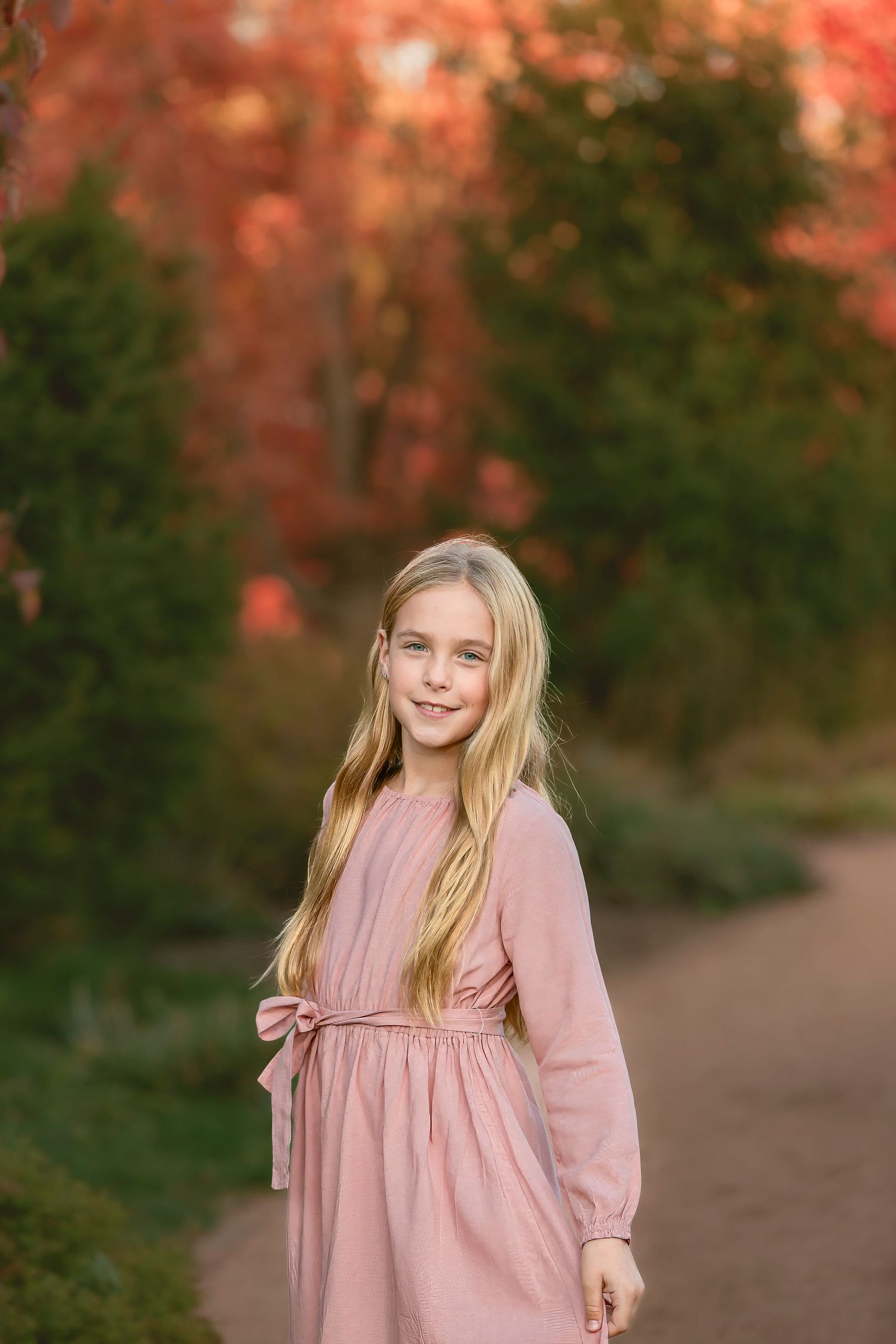A young person in a pink dress stands on a path surrounded by autumn foliage with warm lighting.