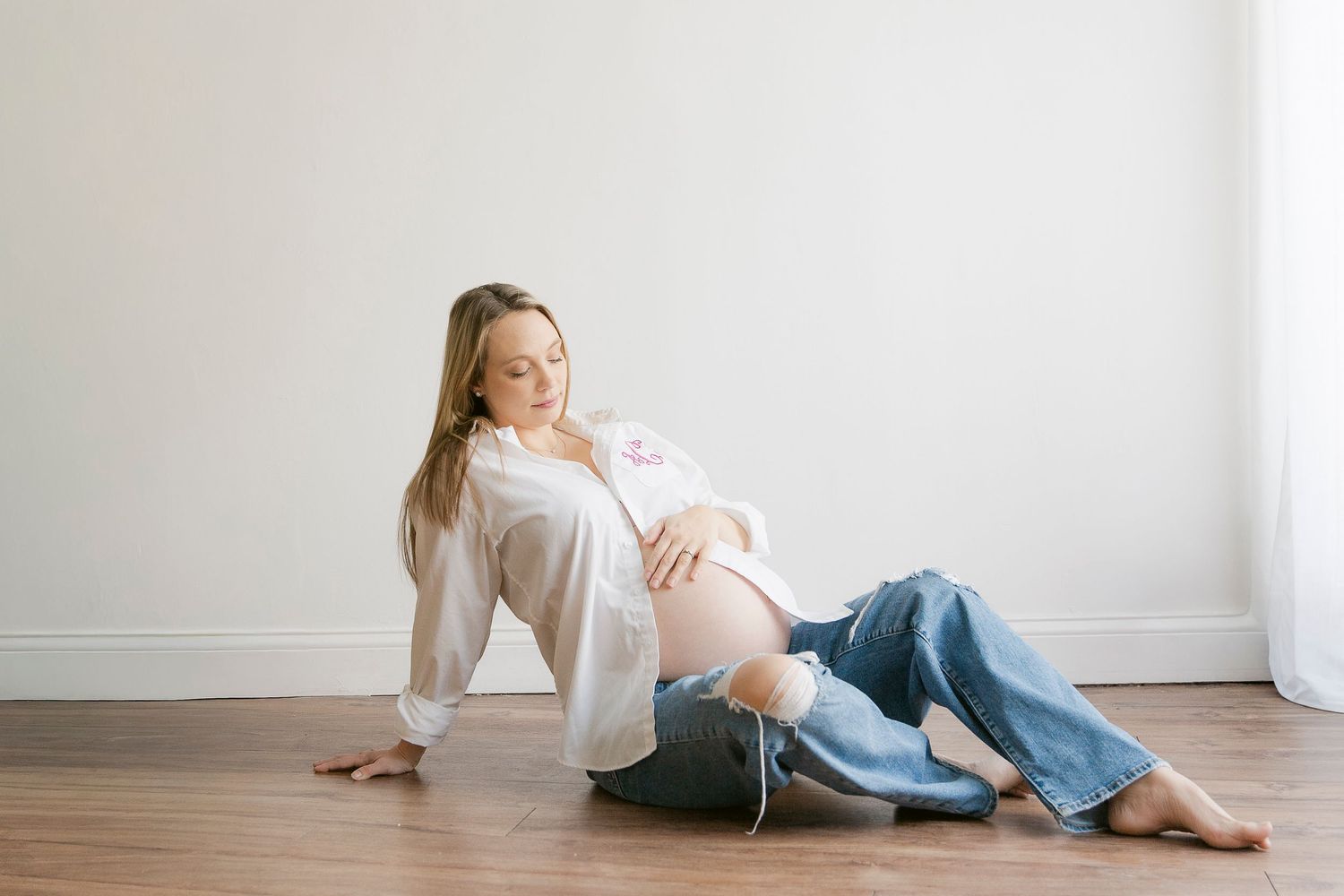 Pregnant woman sitting on hardwood floor in white shirt and ripped jeans, cradling baby bump against white wall.