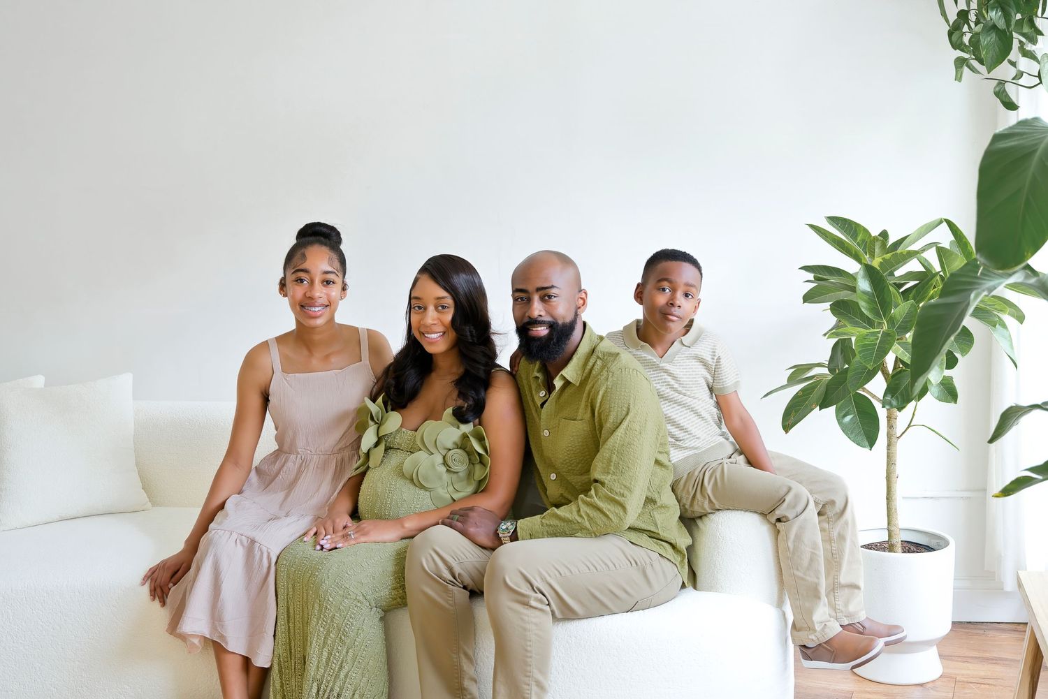 Family of four sitting together on white sofa in bright studio for maternity portrait session.