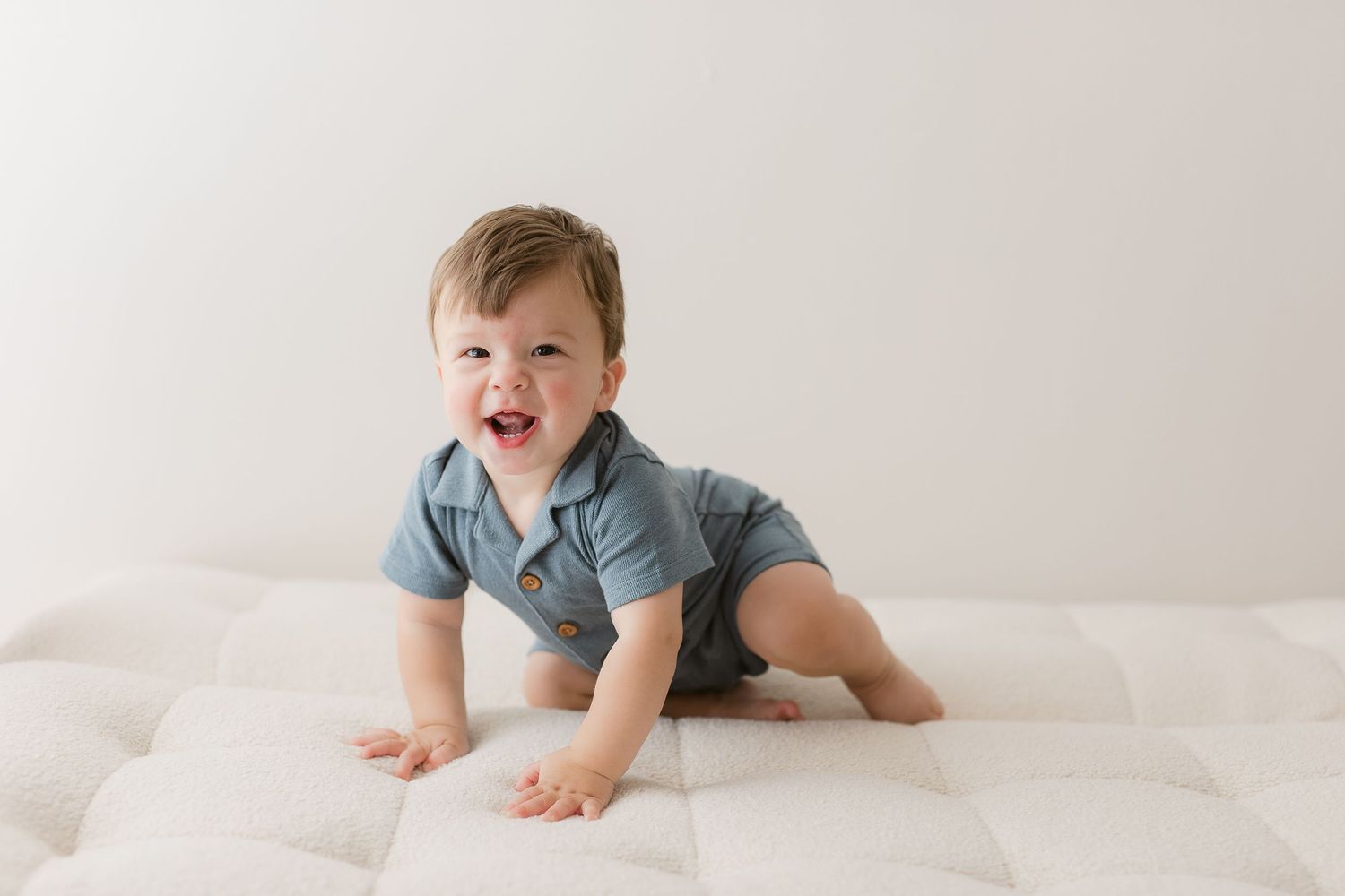 Happy baby in blue onesie crawling on white mattress.
