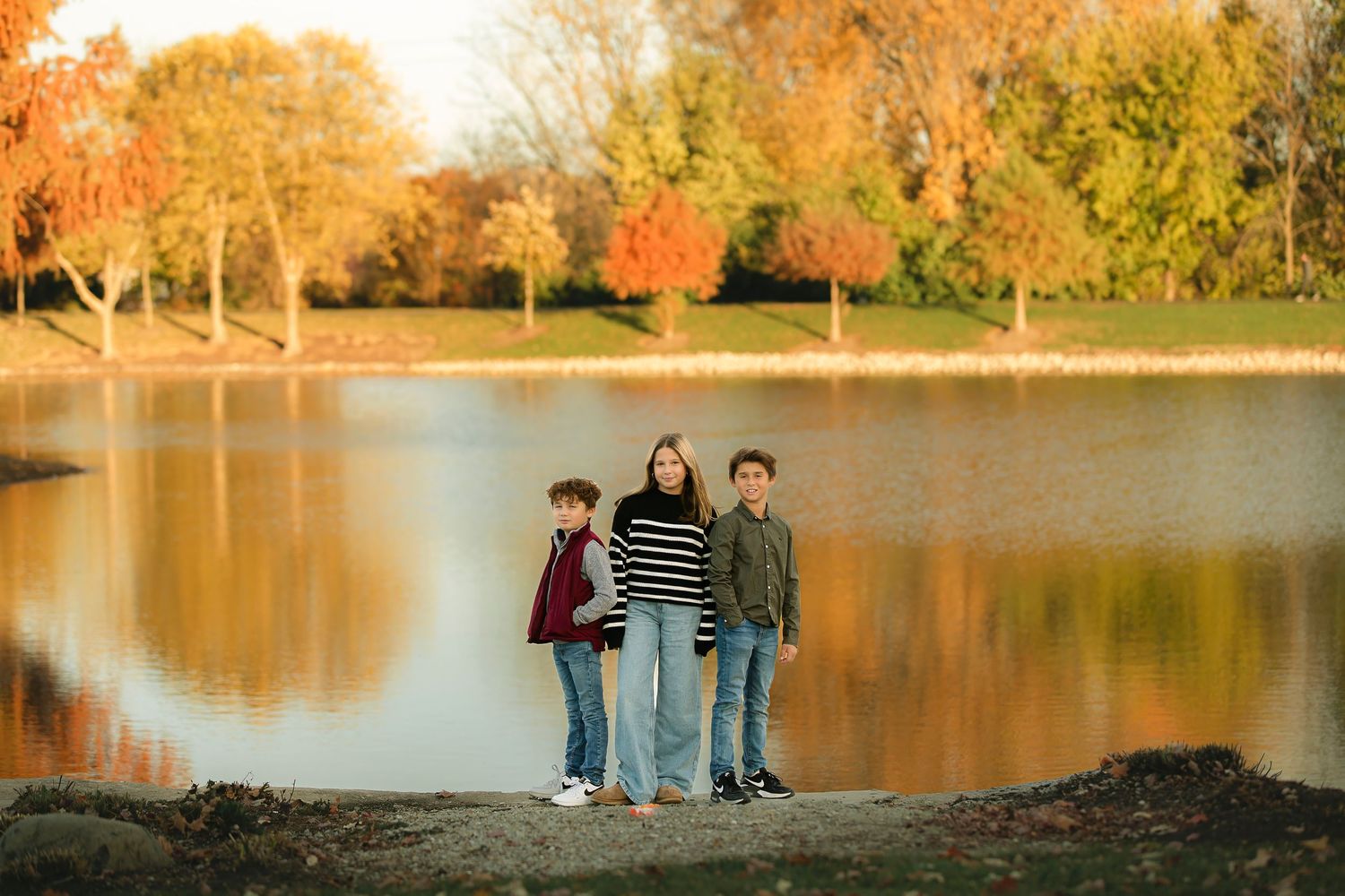 Three people stand together by lake with colorful autumn trees reflecting in water.