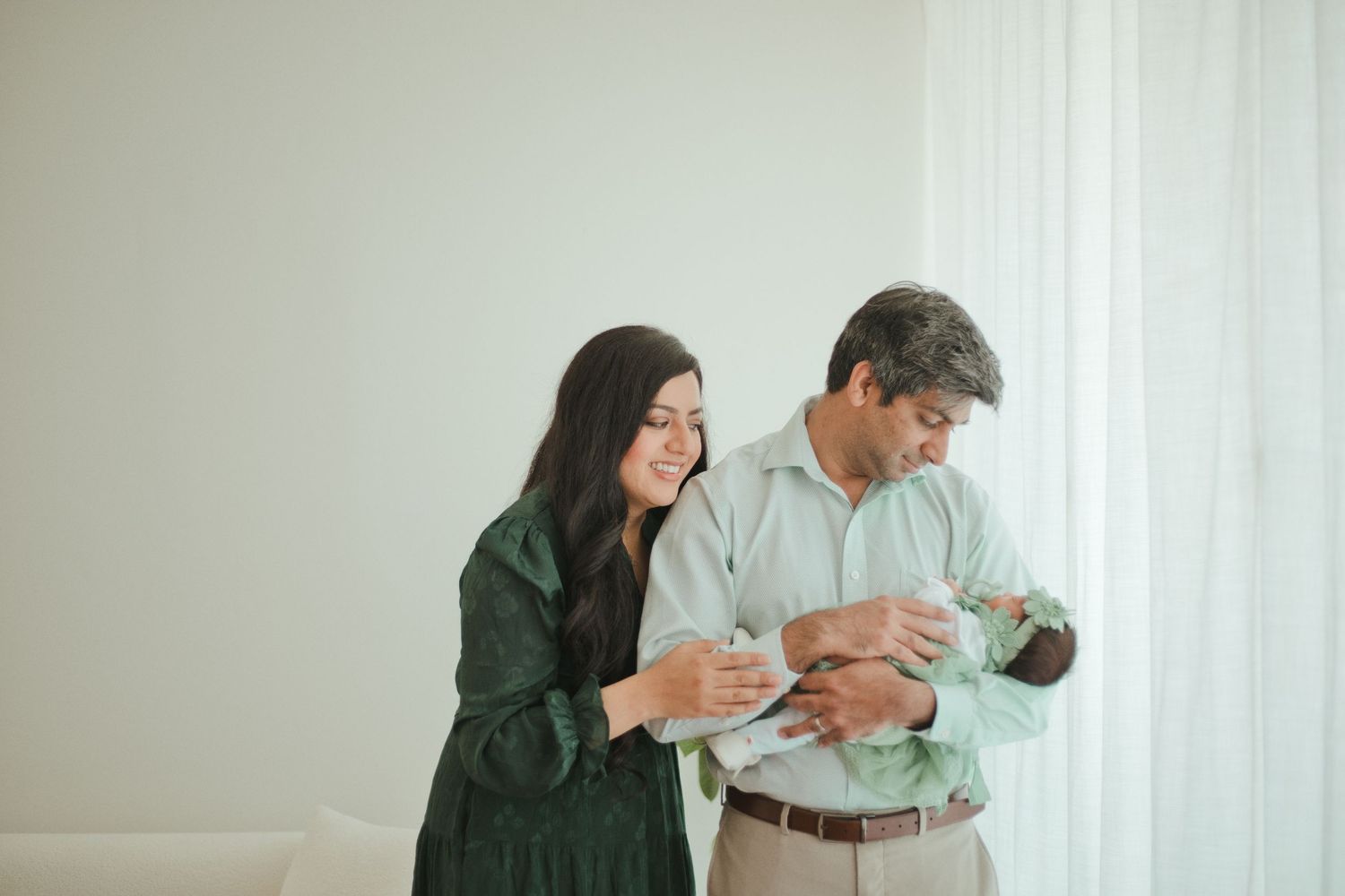 A couple smiles while cradling a newborn baby near a bright window in a softly lit room.