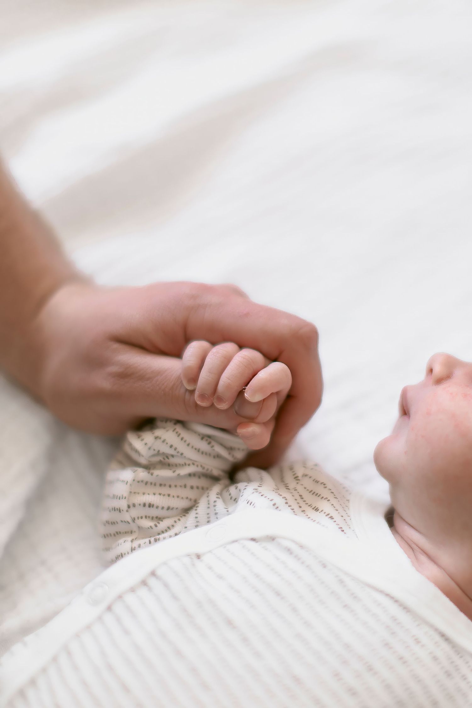 A newborn baby grasping an adult finger while lying on a soft white blanket in a tender moment.