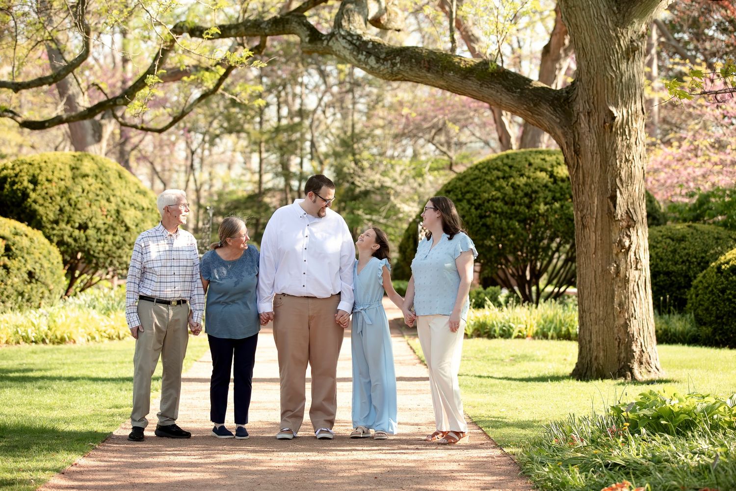 Multi-generational family walking together on a garden path surrounded by lush green trees and manicured shrubs.