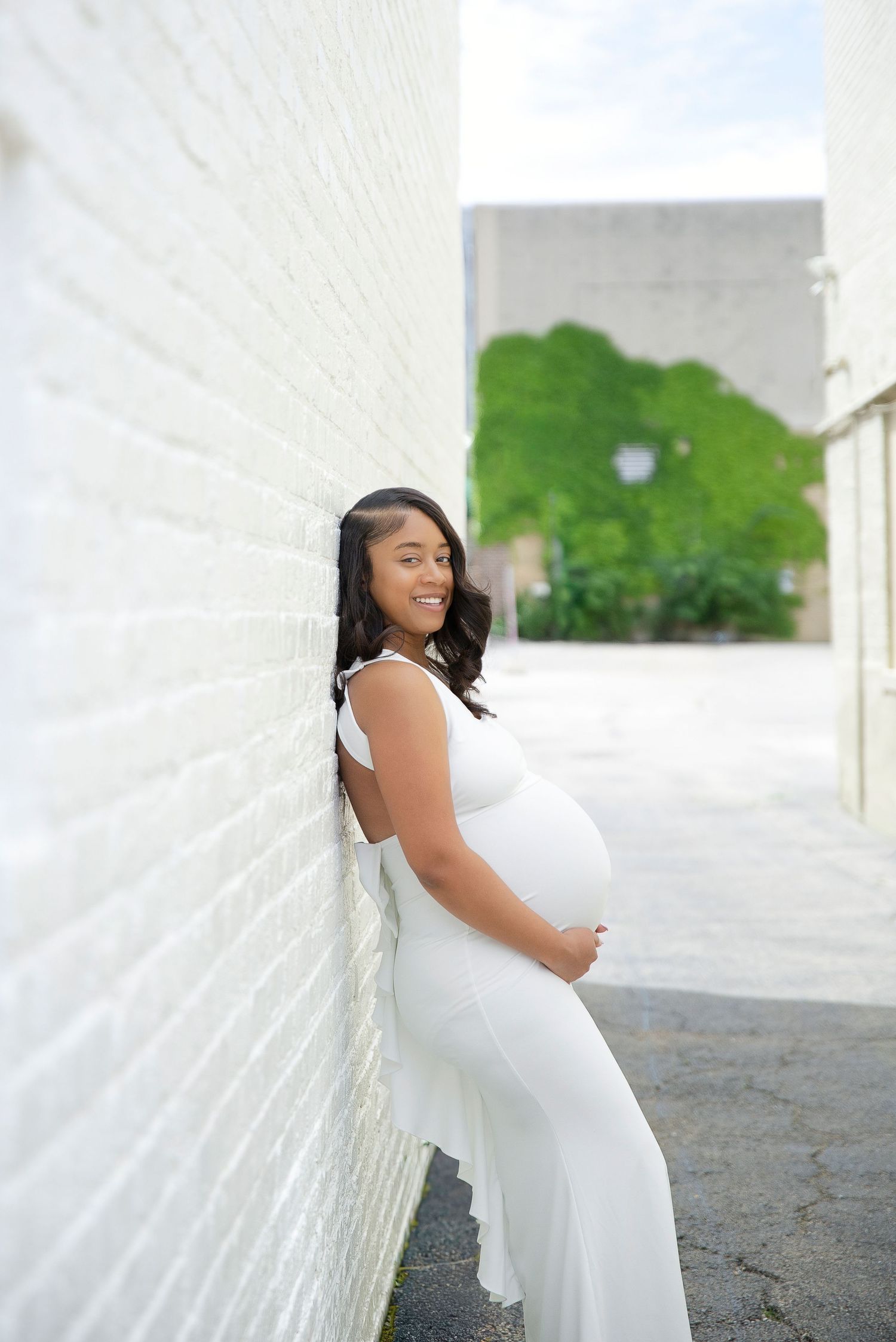 Smiling pregnant woman in white jumpsuit leans against white brick wall outdoors.