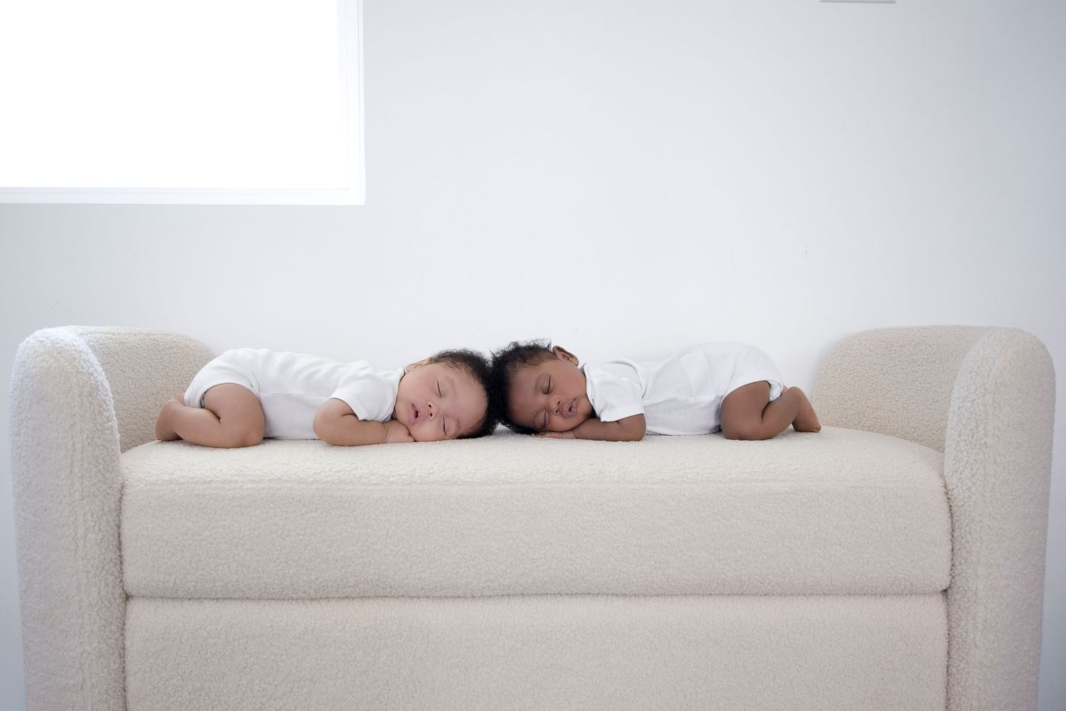 Two babies in white onesies sleeping side by side on a cream sofa in a bright white room.