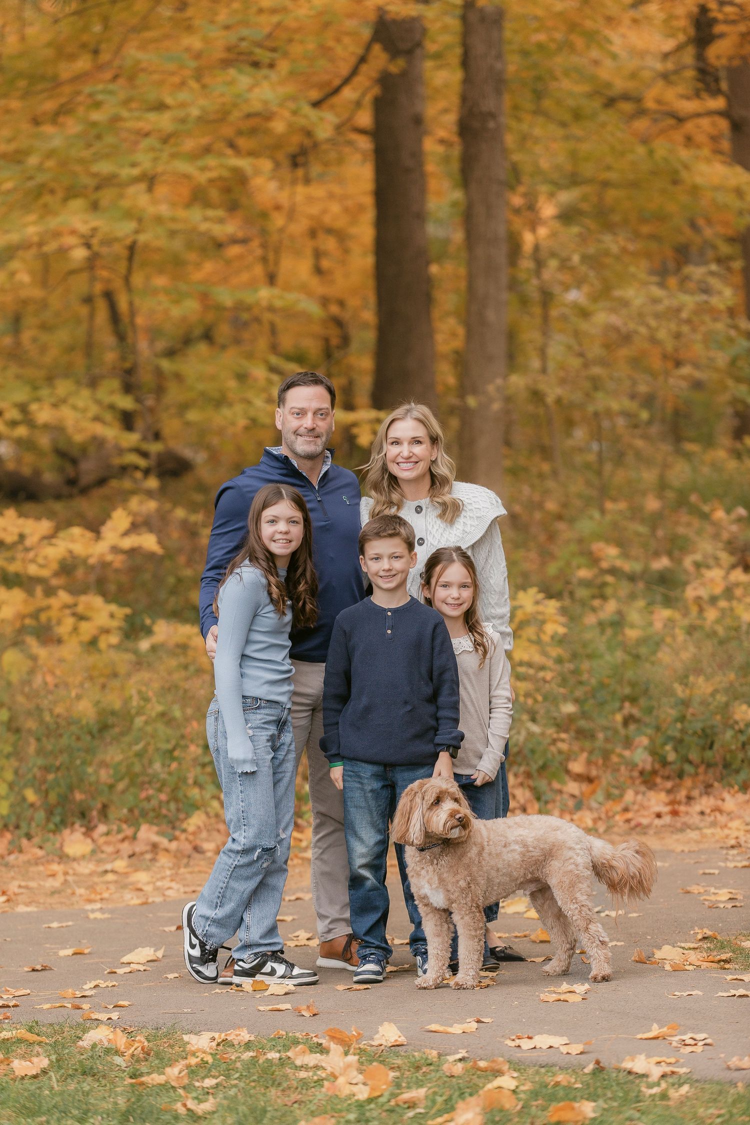 A family and their fluffy brown dog pose together on a path surrounded by golden autumn foliage in a park.
