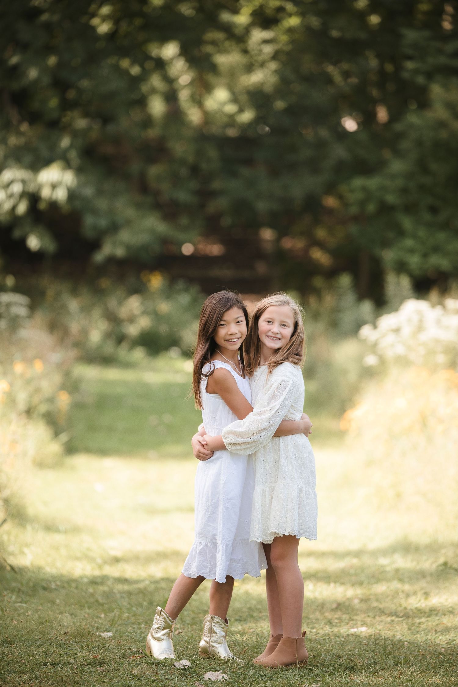 Two young girls in white dresses hugging and smiling outdoors in a lush green garden setting.