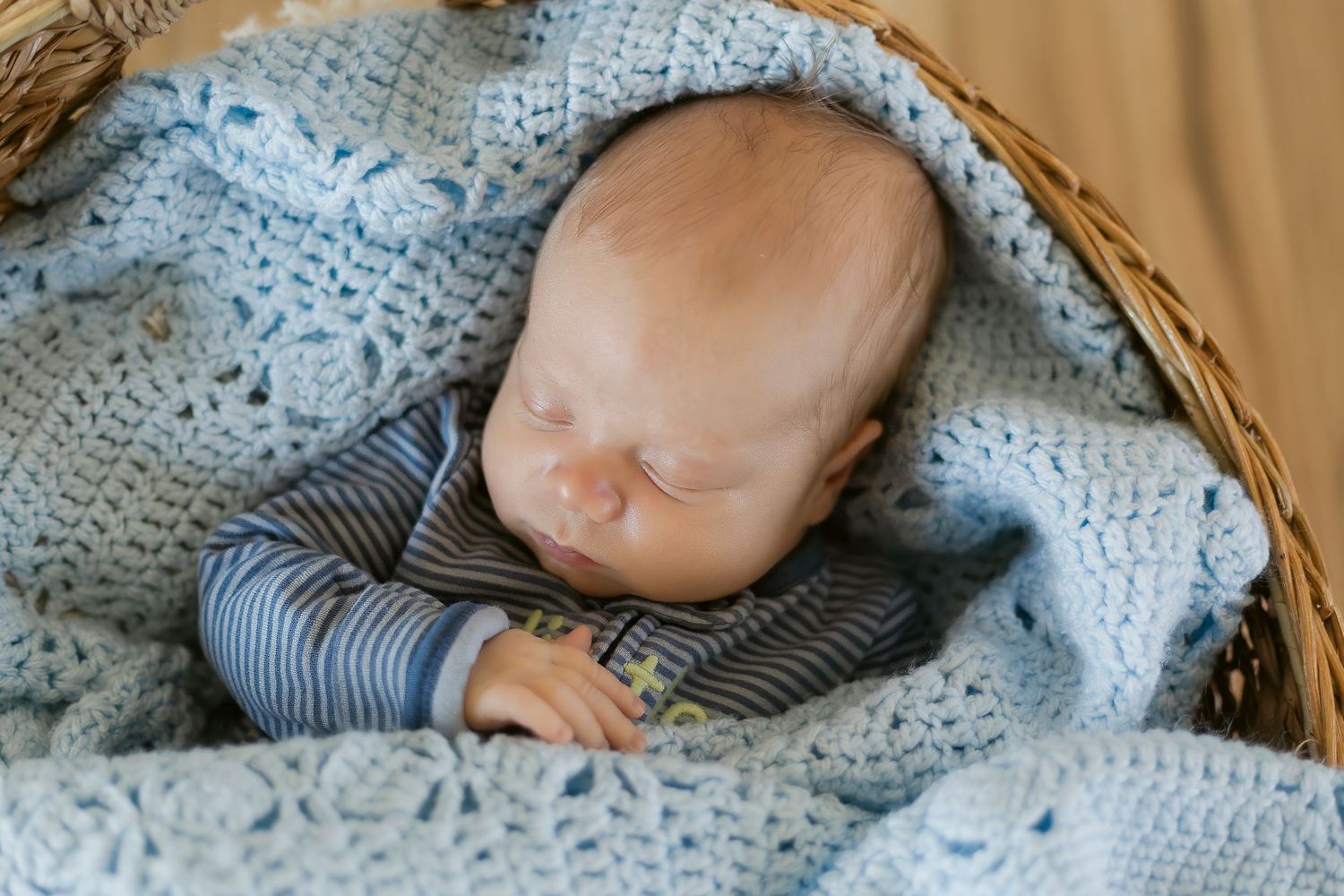 Newborn baby sleeping peacefully in a wicker basket wrapped in a soft blue crochet blanket.