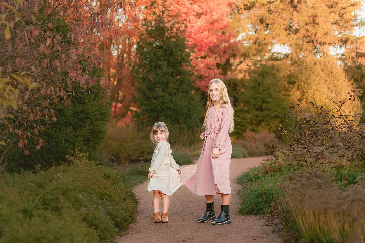 Two children in pink dresses walk along an autumn path surrounded by colorful fall foliage at sunset.