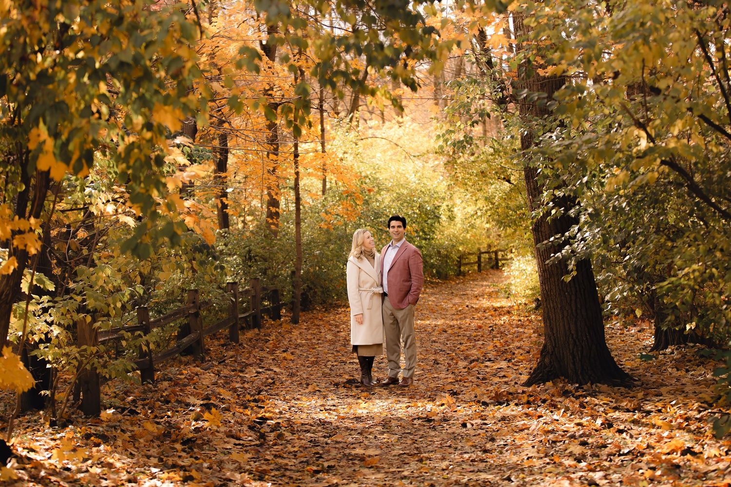 A couple walks hand in hand down a tree-lined autumn path covered in golden fallen leaves.