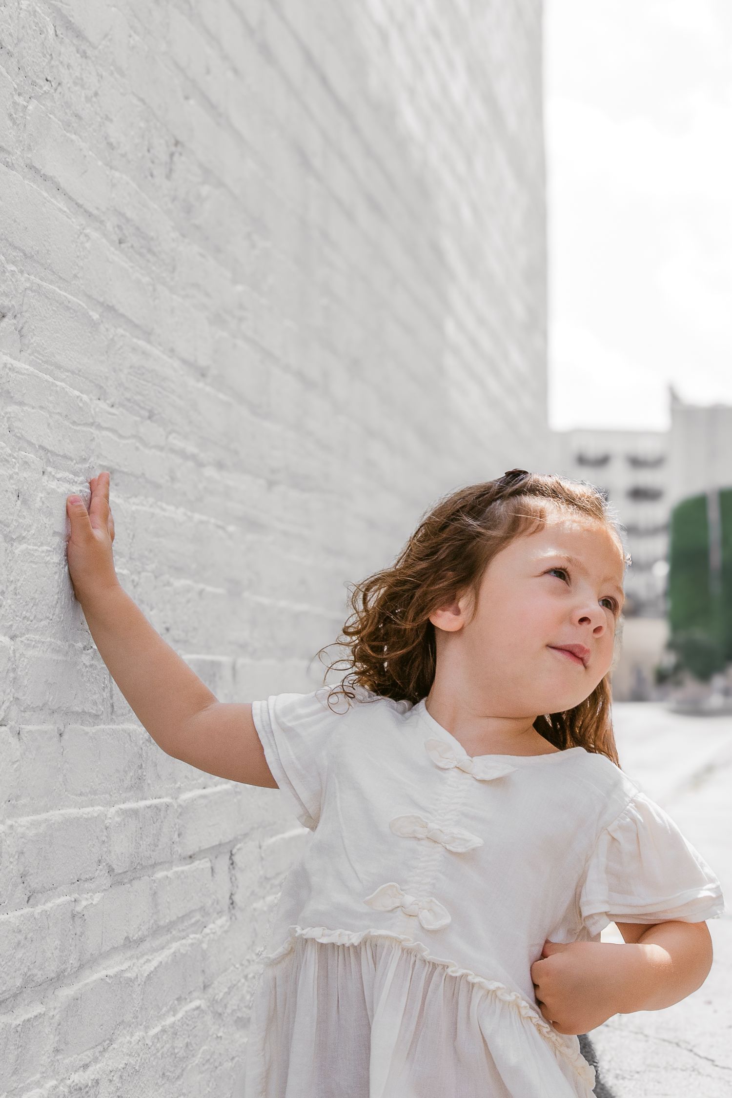 Young child in white dress touching white brick wall while looking up with curiosity.