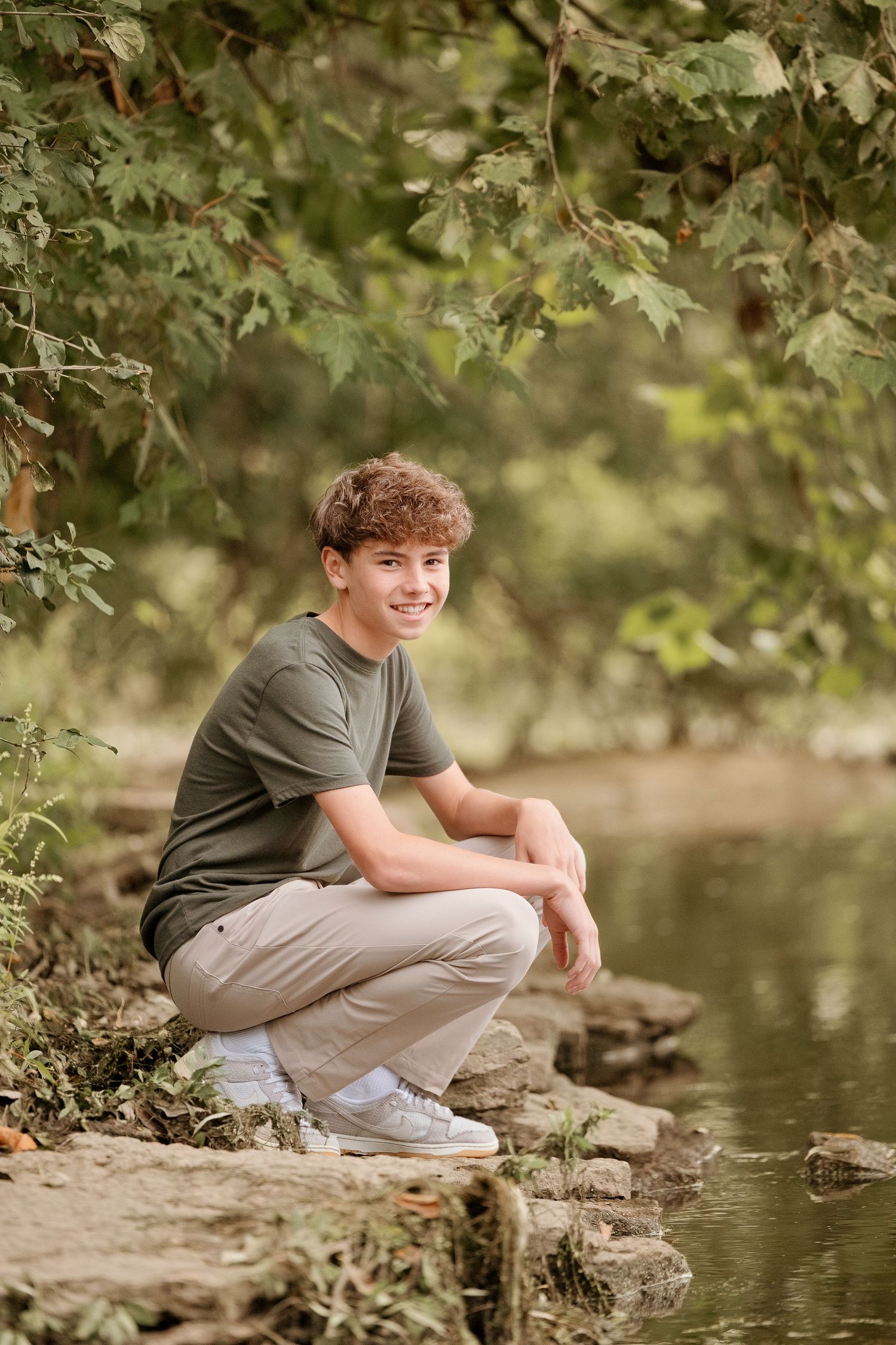 A smiling teenage boy crouches on rocks by a creek, wearing a olive green t-shirt and khaki pants in a lush forest.