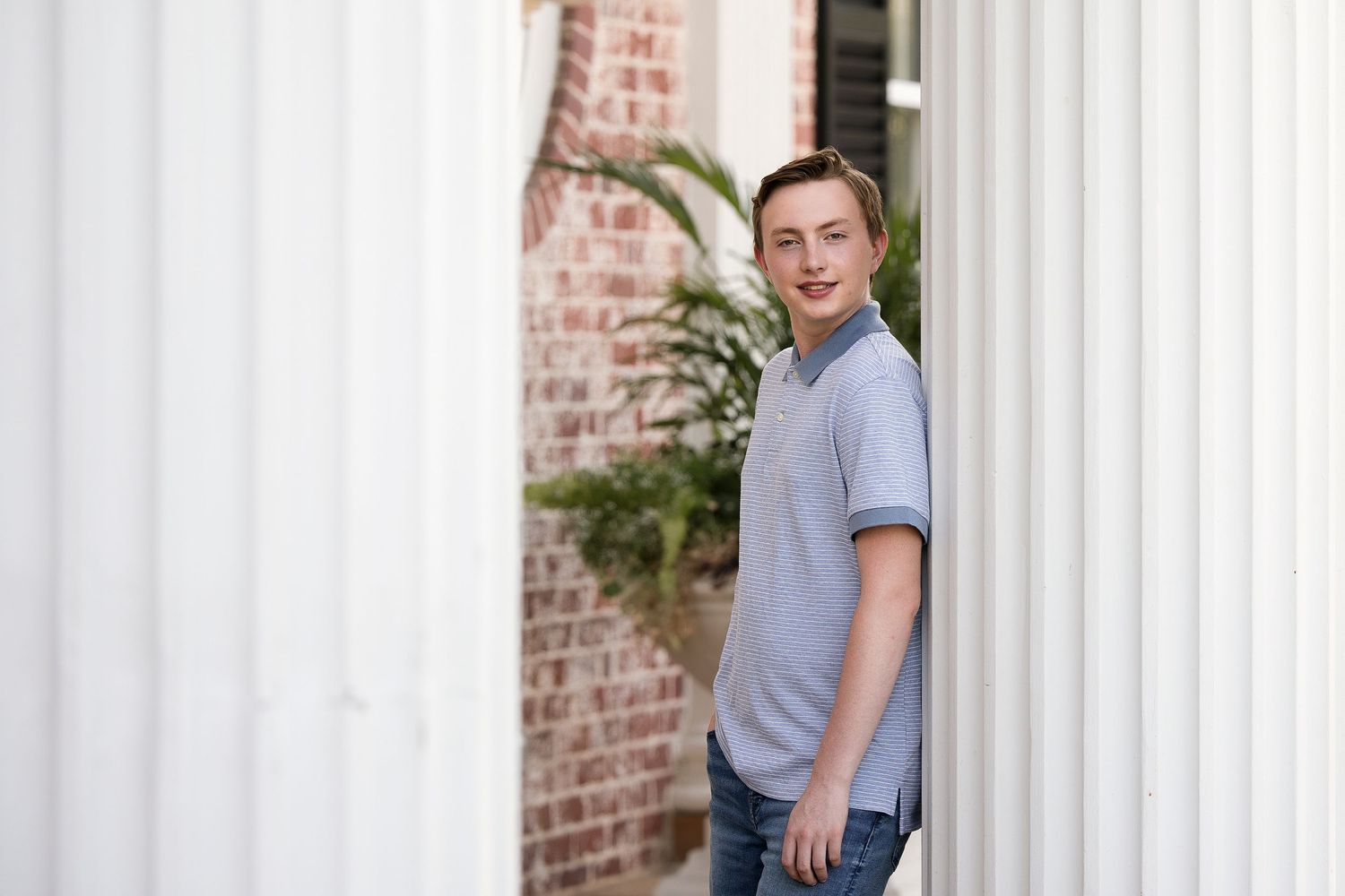 Teen boy in gray polo shirt leaning against white columns near brick wall for senior portrait.