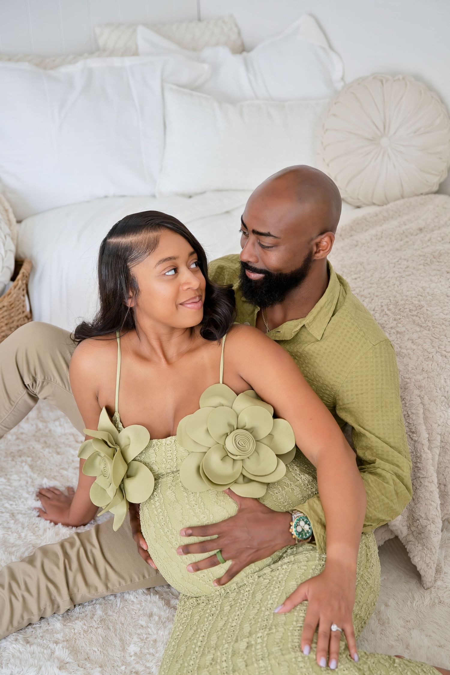 A pregnant woman in a green floral dress sits with her partner on a bedroom floor, both smiling warmly.