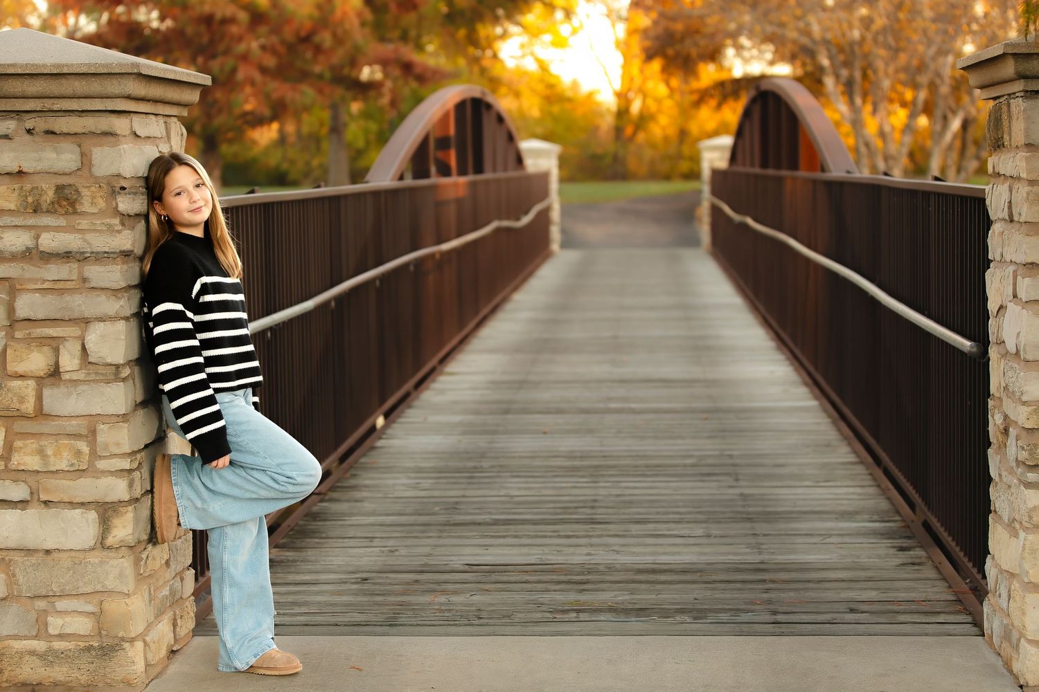 Person in striped shirt and jeans poses on wooden bridge during golden autumn sunset.