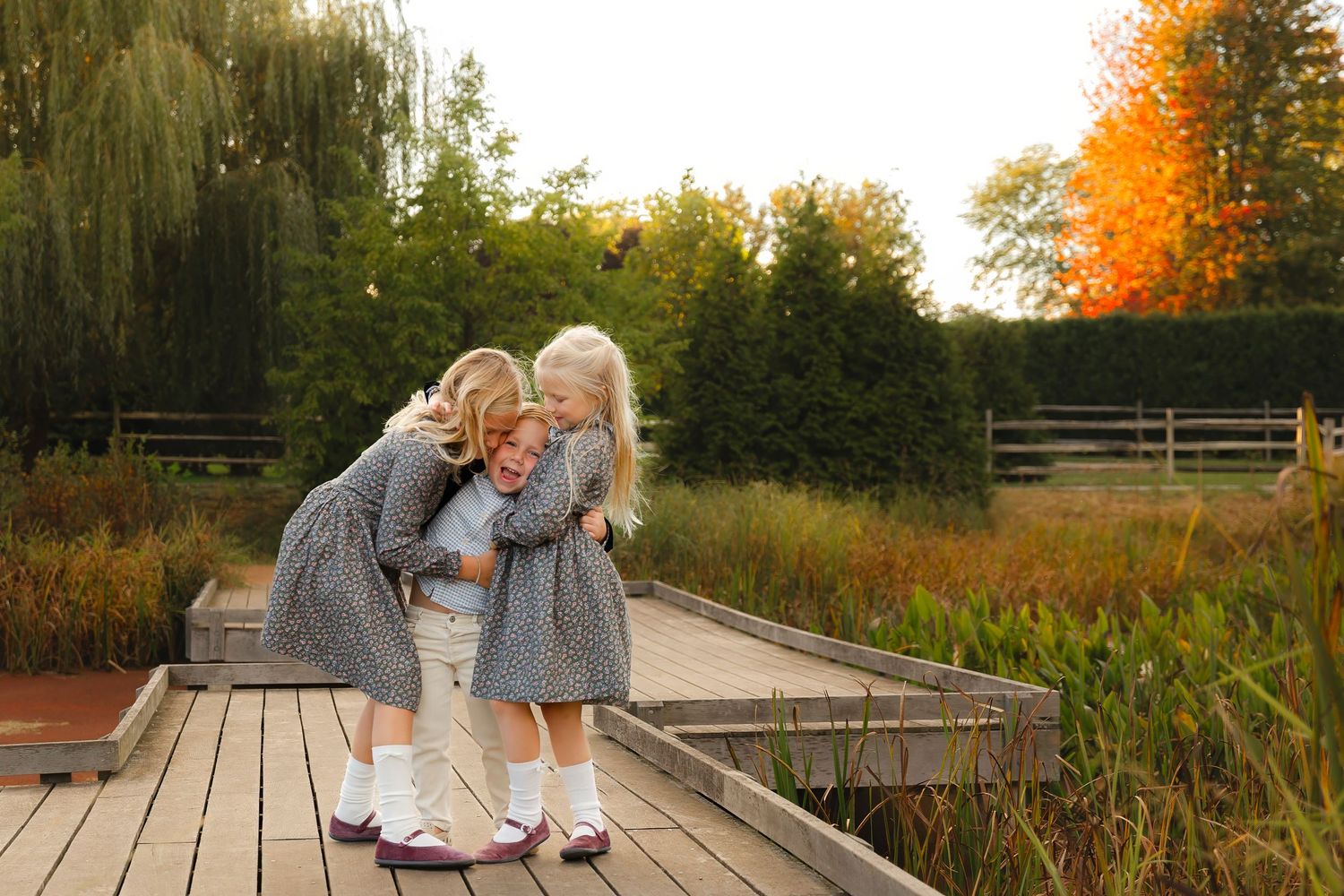 Two children in gray dresses share a sweet moment on a wooden boardwalk during autumn.