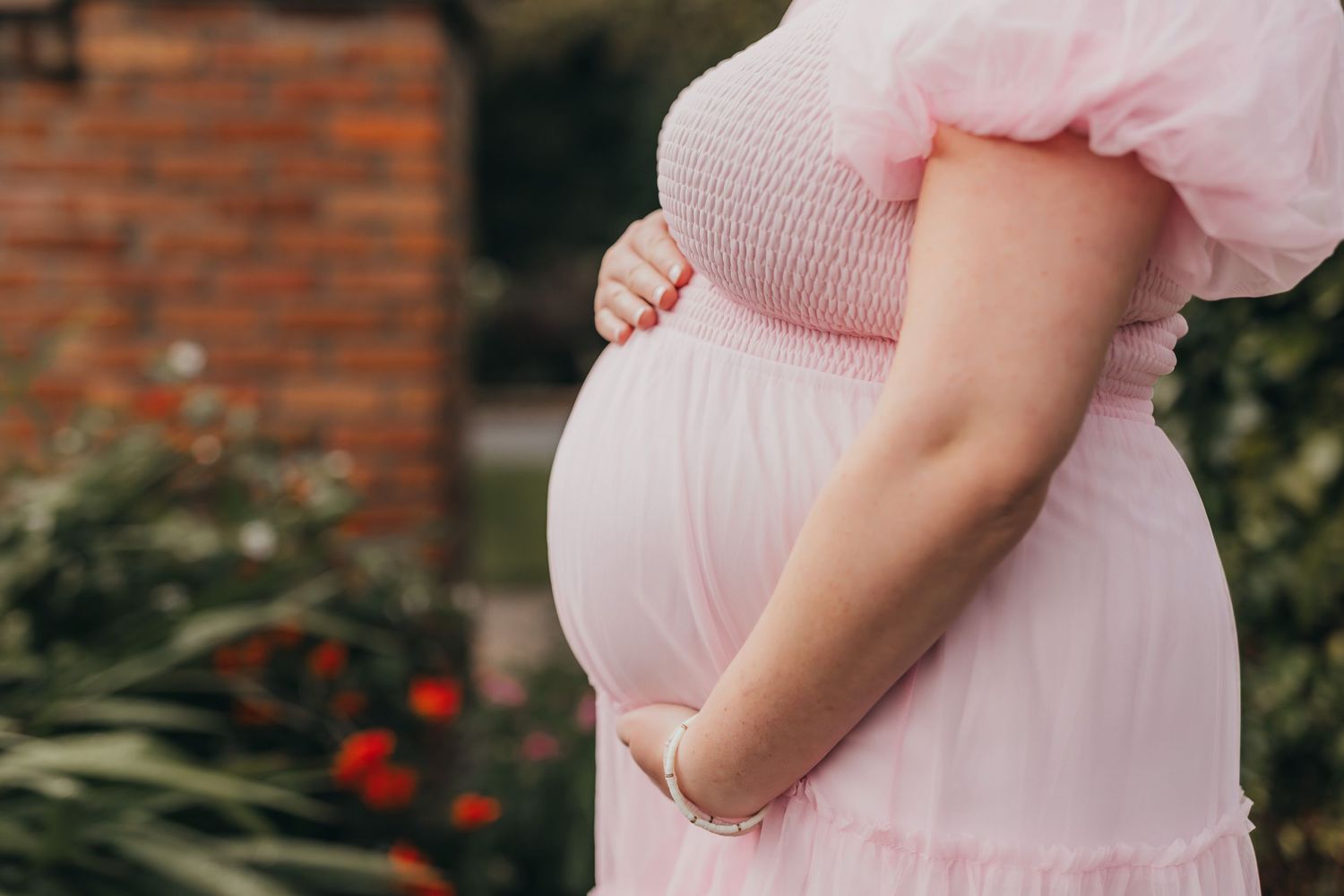 Side view of pregnant woman in pink dress touching her baby bump in garden setting.