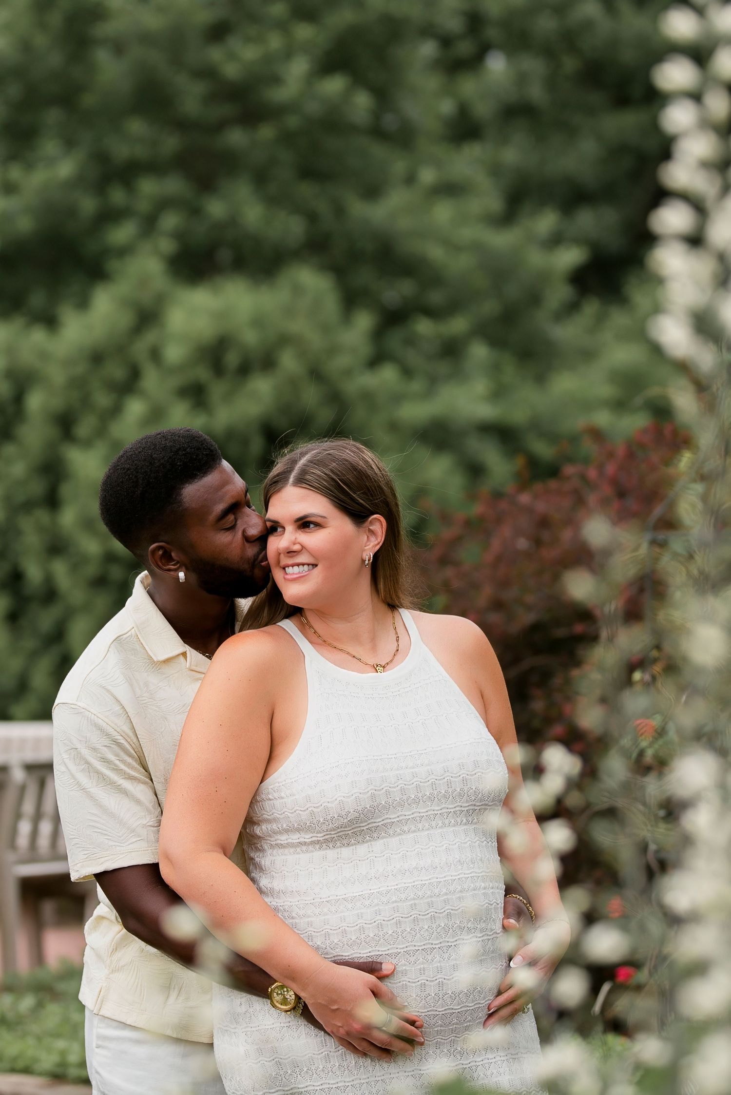 A couple shares a tender moment outdoors, the woman in a white dress cradling her baby bump while her partner kisses her cheek.