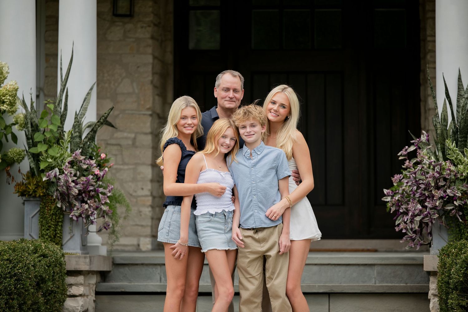 A happy family of five poses together on the front steps of an elegant stone home surrounded by lush greenery.