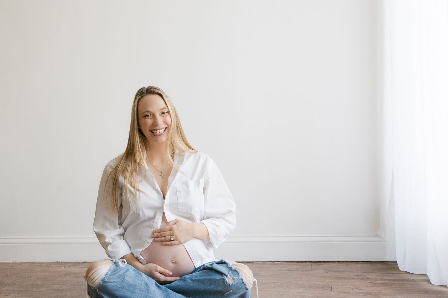 Woman in white shirt sitting on floor against white wall giving thumbs up and smiling.