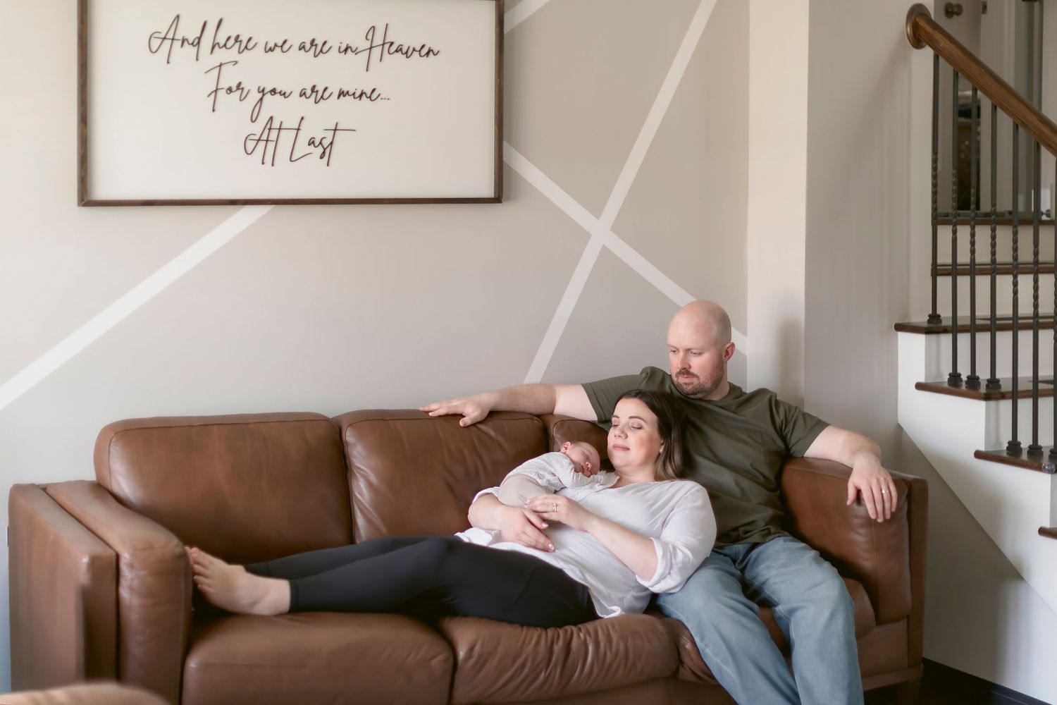 Couple relaxing on brown leather sofa with newborn baby in cozy home with inspirational wall sign.