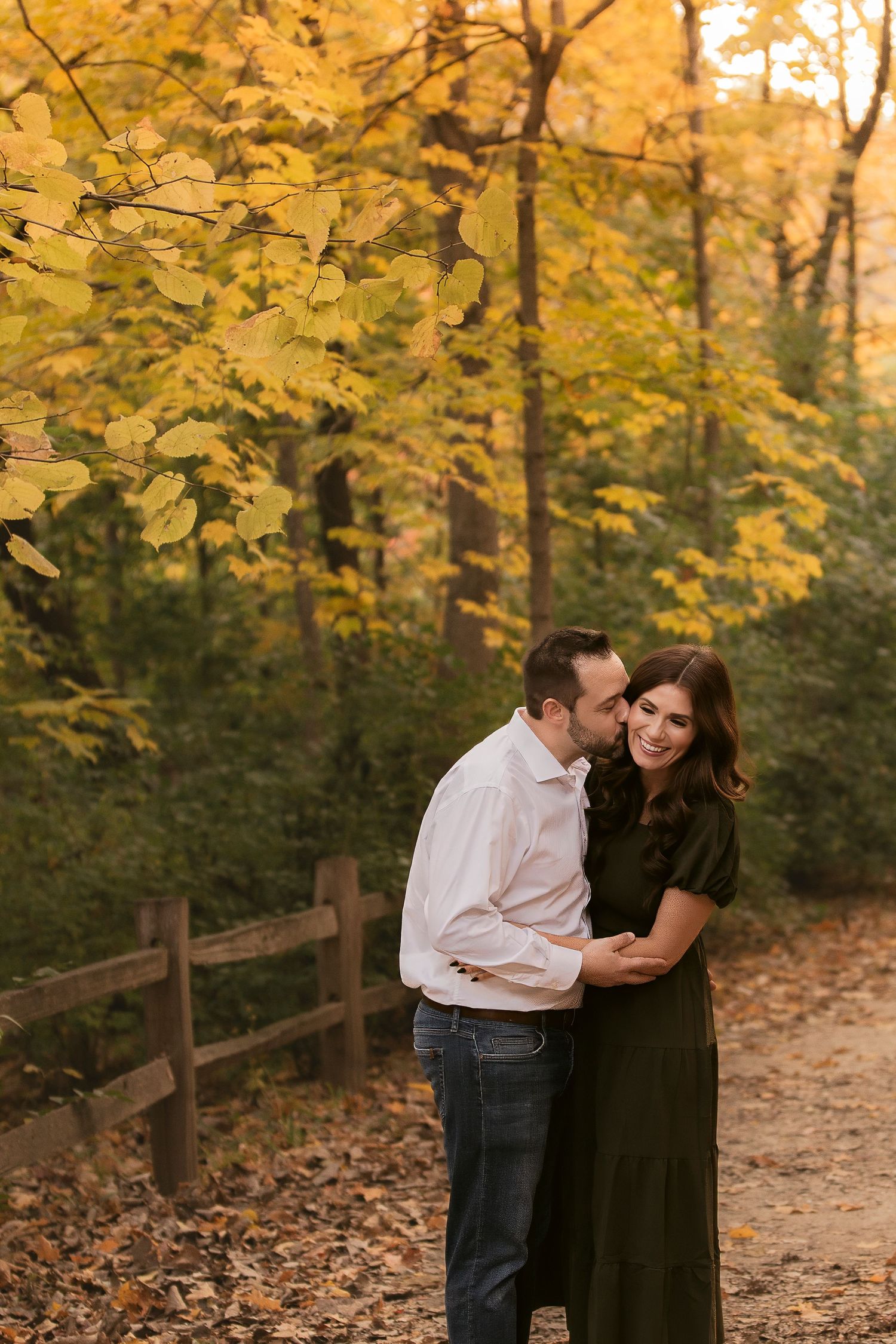 Couple sharing a romantic kiss among vibrant autumn foliage on a wooded path during golden hour.