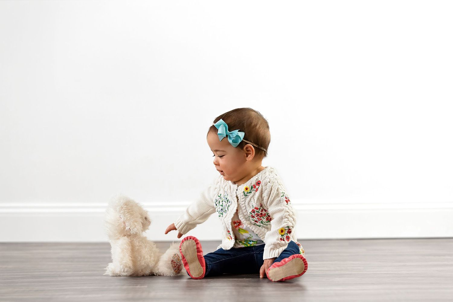 Small child wearing sunglasses sits on hardwood floor playing with a plush teddy bear toy.