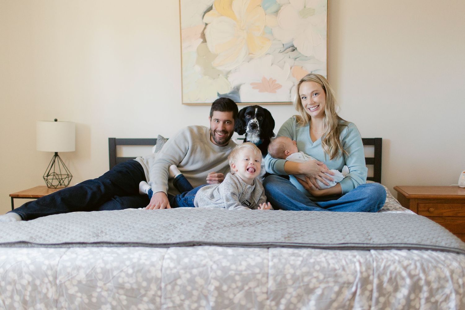 Family with toddler, newborn, and dog smiling together on a bed in a cozy bedroom setting.